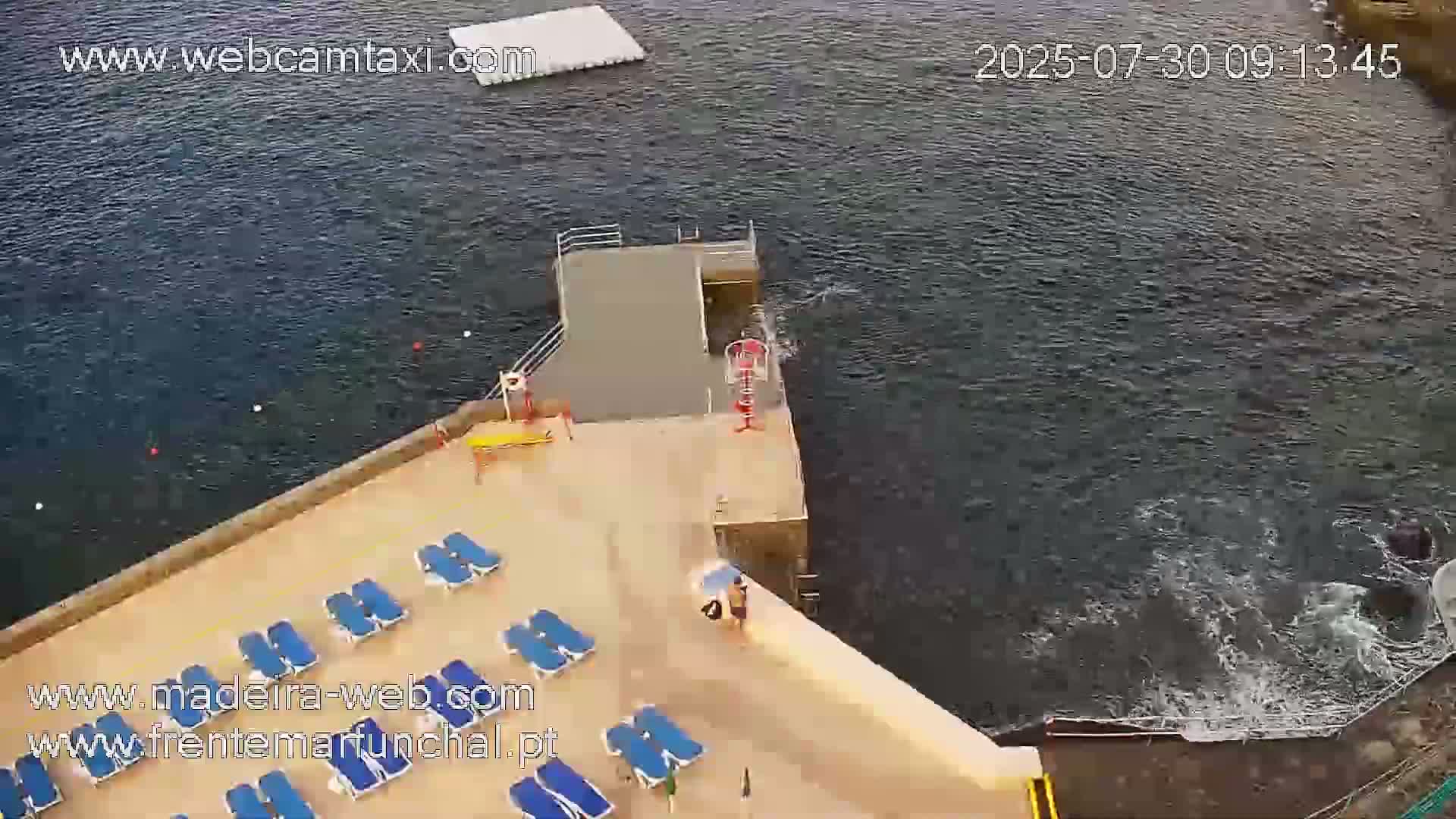 A tan-colored pool deck with blue lounge chairs next to dark-blue water, on a mostly sunny day.