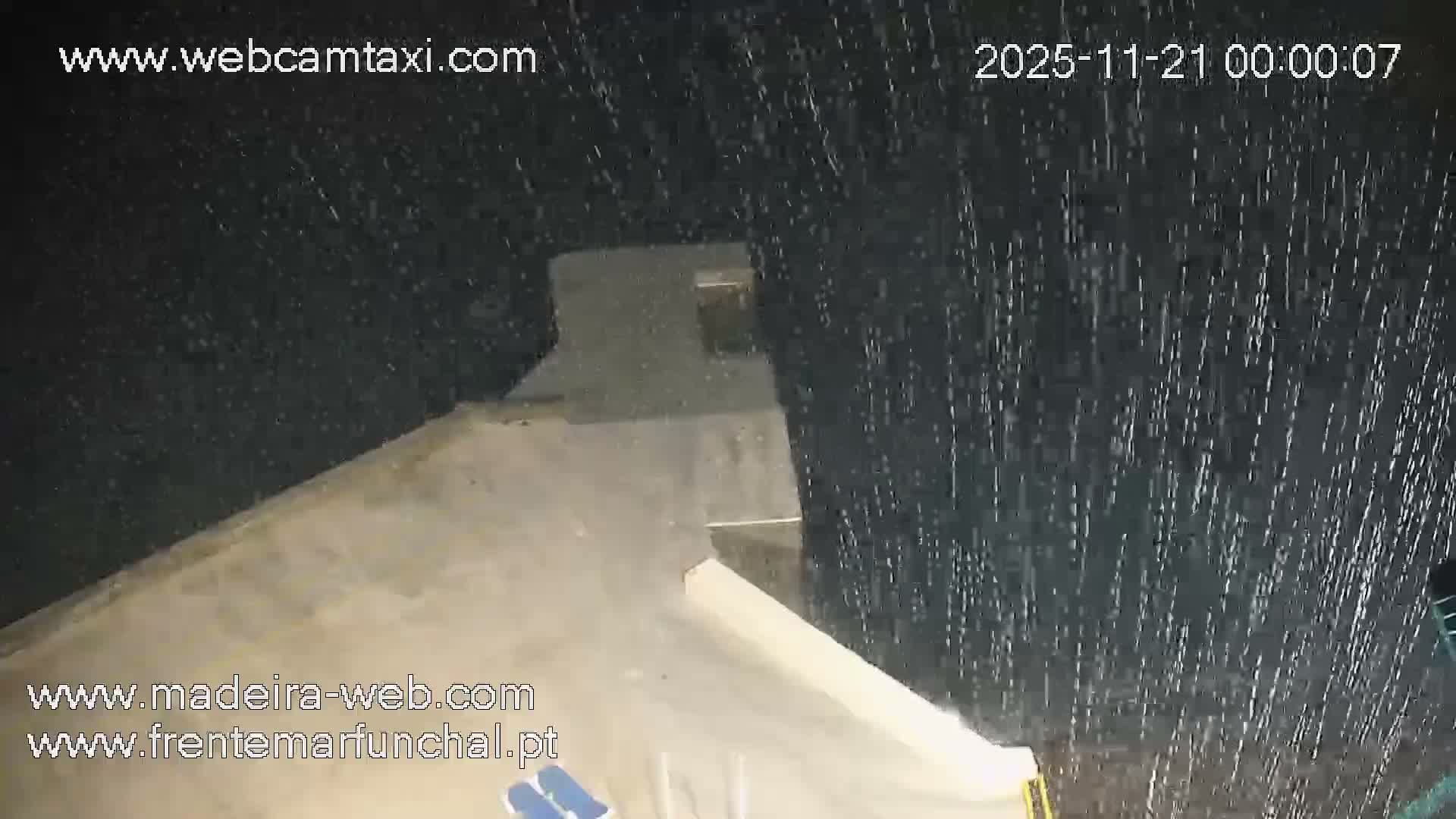 A nighttime view shows a concrete patio with empty lounge chairs next to a dark body of water with small waves lapping against a low seawall.