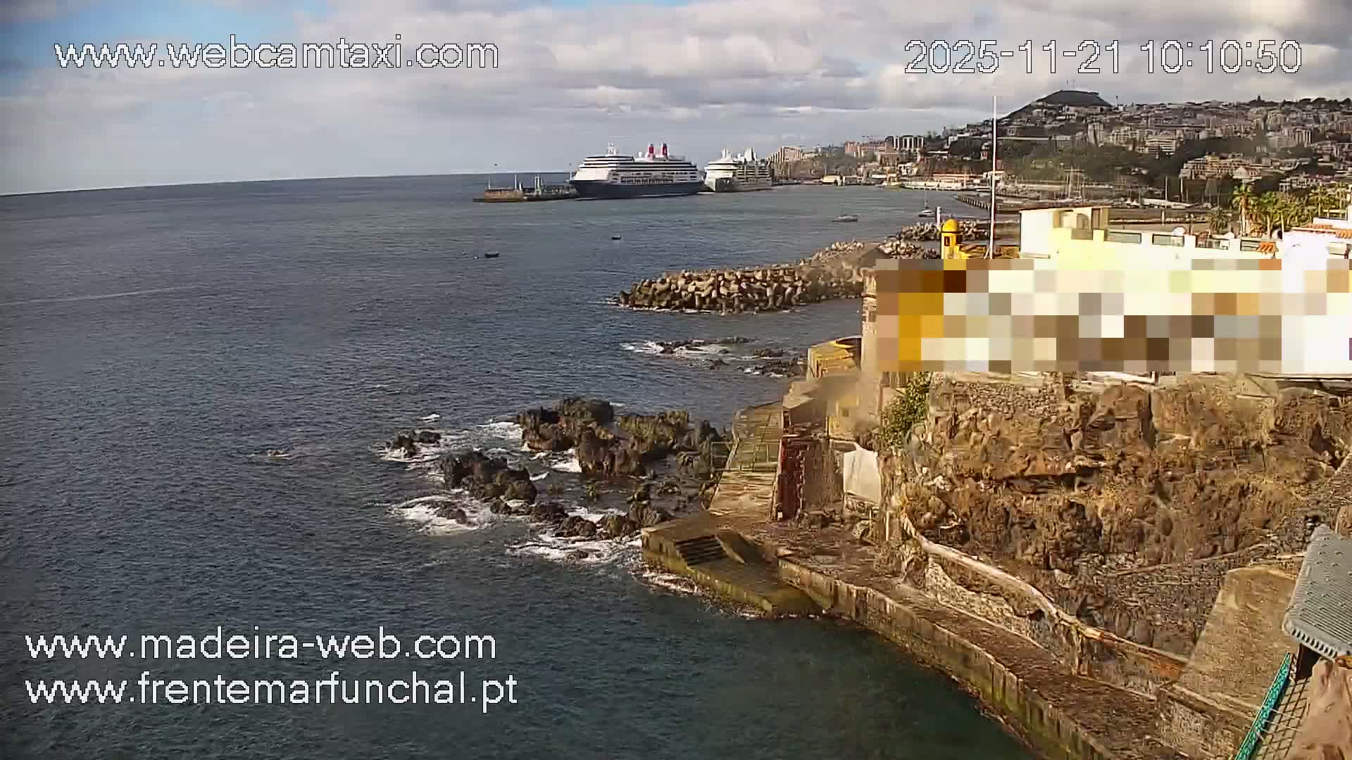 A coastal city built into hills overlooks a harbor where two large cruise ships are docked alongside a rocky shoreline and breakwater, all bathed in sunlight under a partly cloudy sky.