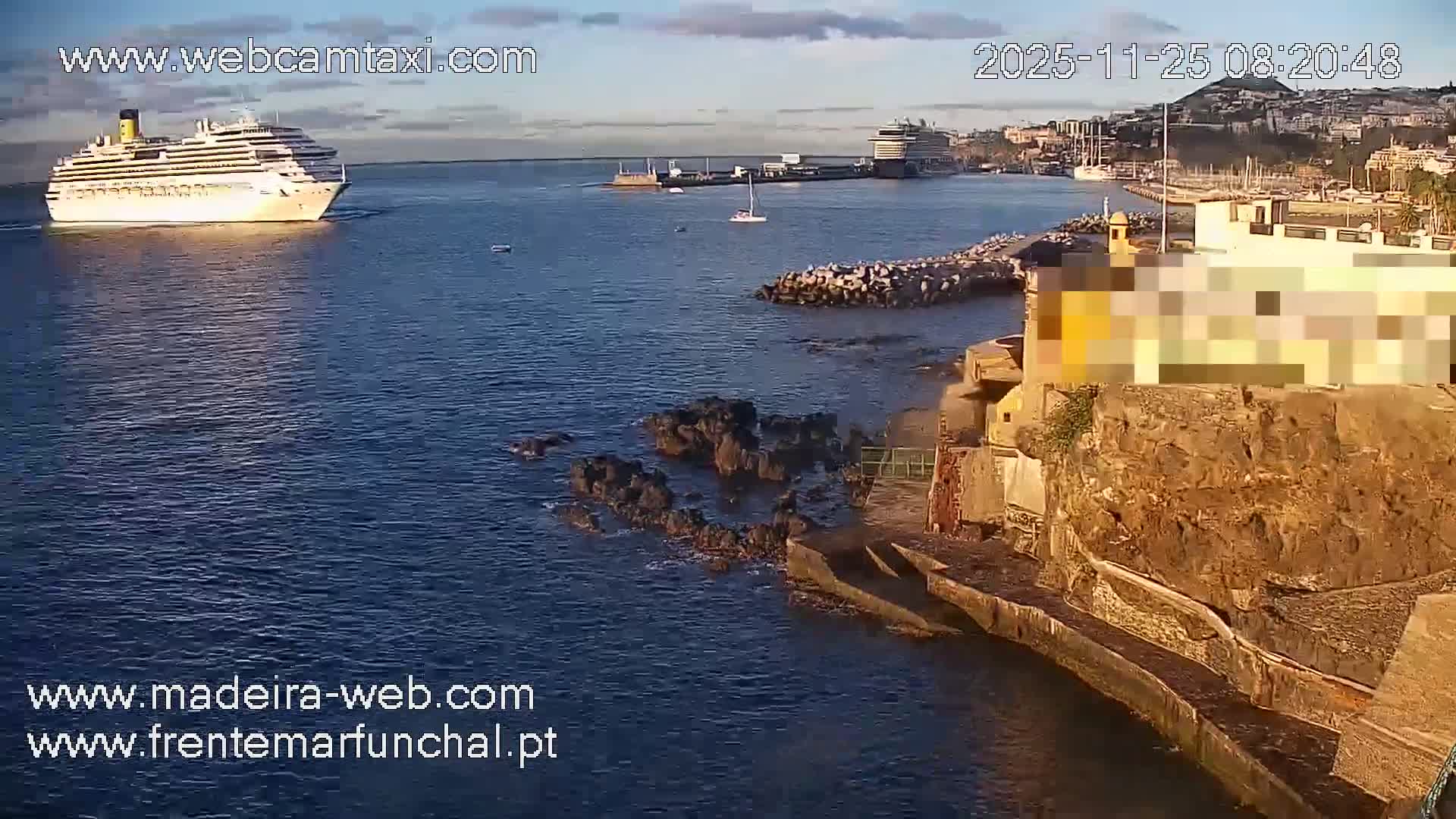 A large white cruise ship sails out of a harbor with calm blue waters under a partly cloudy sky, as another cruise ship remains docked and a coastal city stretches into the background.