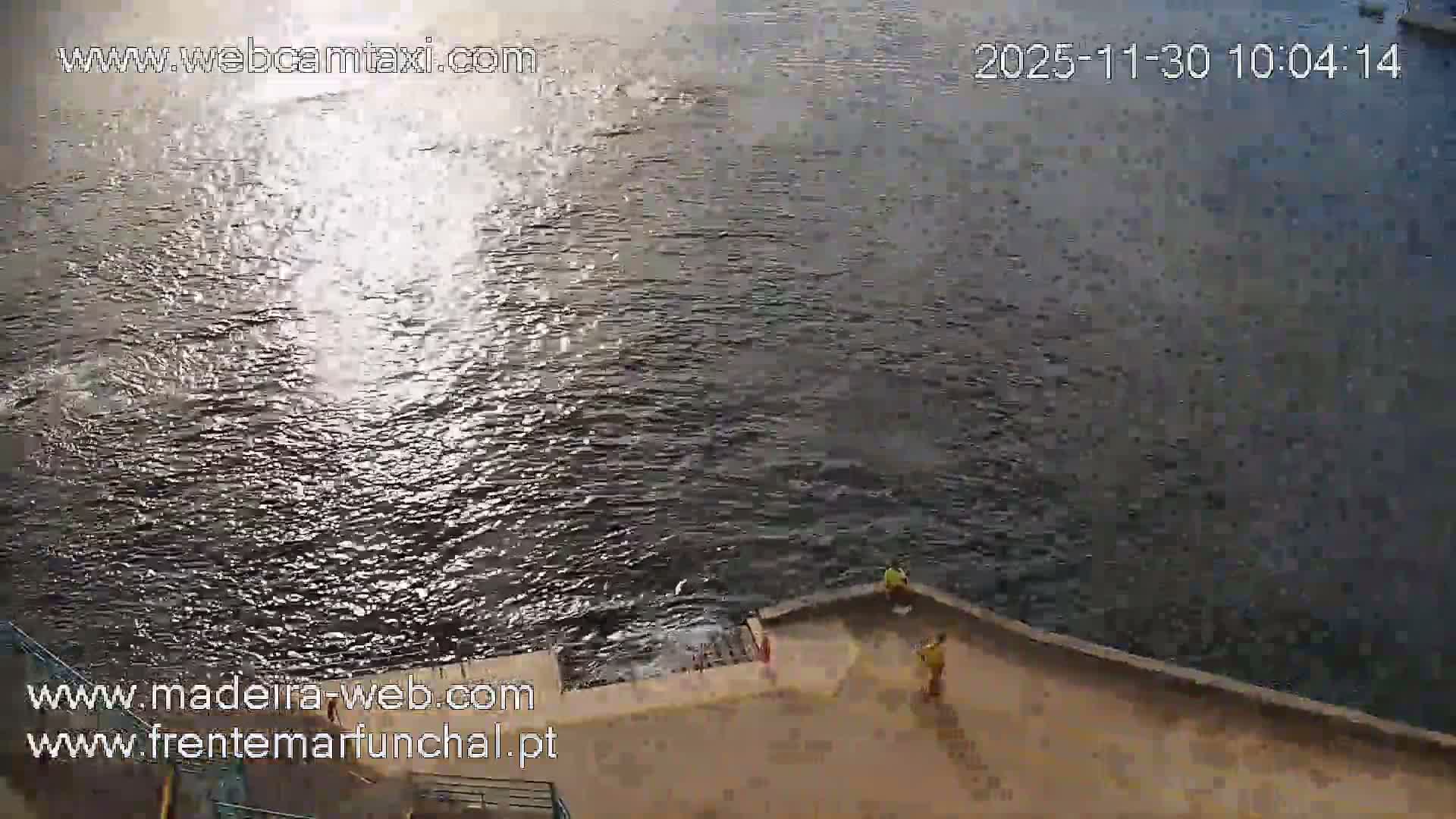 Two people stand on a concrete pier adjacent to a body of choppy water where bright sunlight reflects, indicating a sunny day.