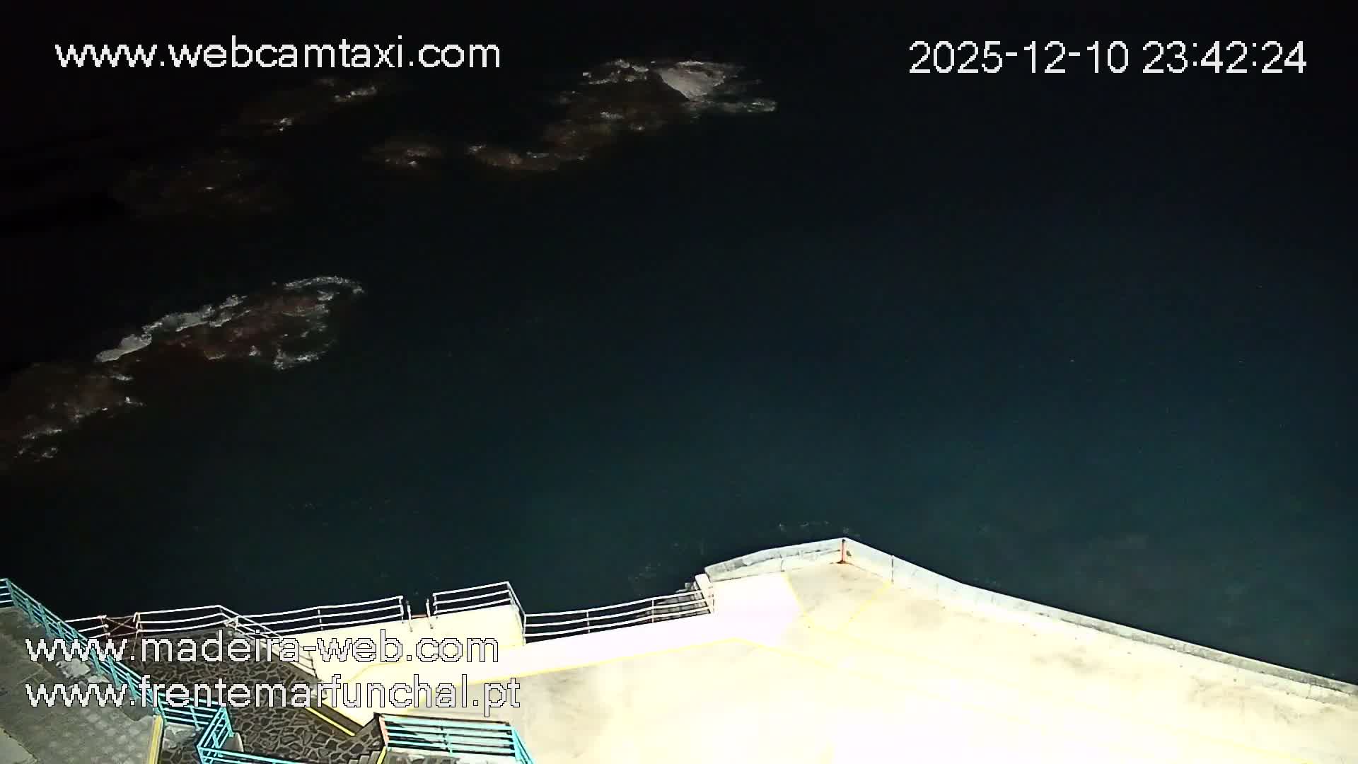 The image displays a dark body of water, likely the sea, reflecting faint lights under a partly cloudy night sky, with a concrete pier and railings visible in the foreground.
