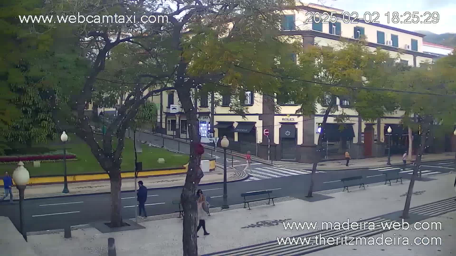 A street scene shows a pedestrian area with benches, a crosswalk, and several people walking past a multi-story building on an overcast day.