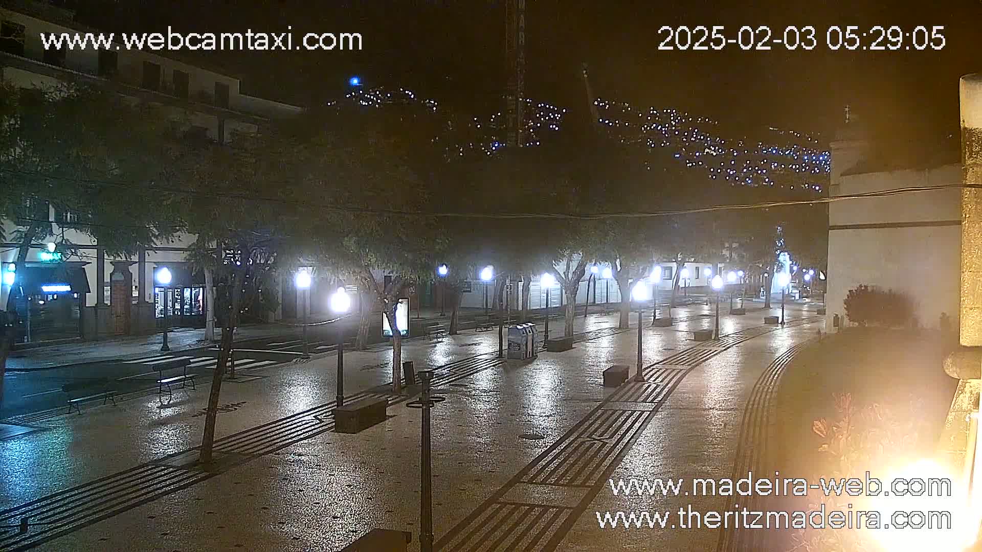 A wet, nighttime town square is lit by streetlights, with buildings and hills visible in the background.