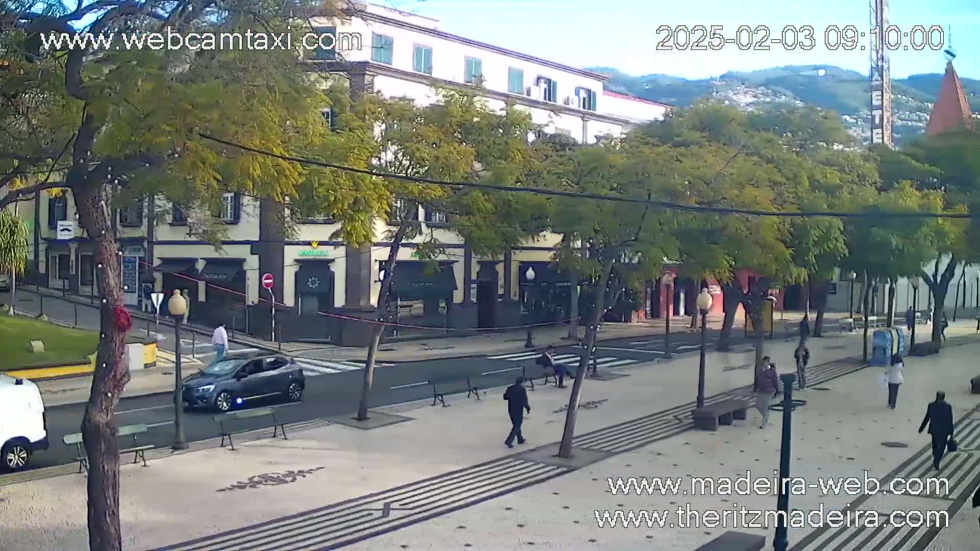 A street scene on a sunny day shows a pedestrian area with people walking past shops and buildings, and a car driving down the road.