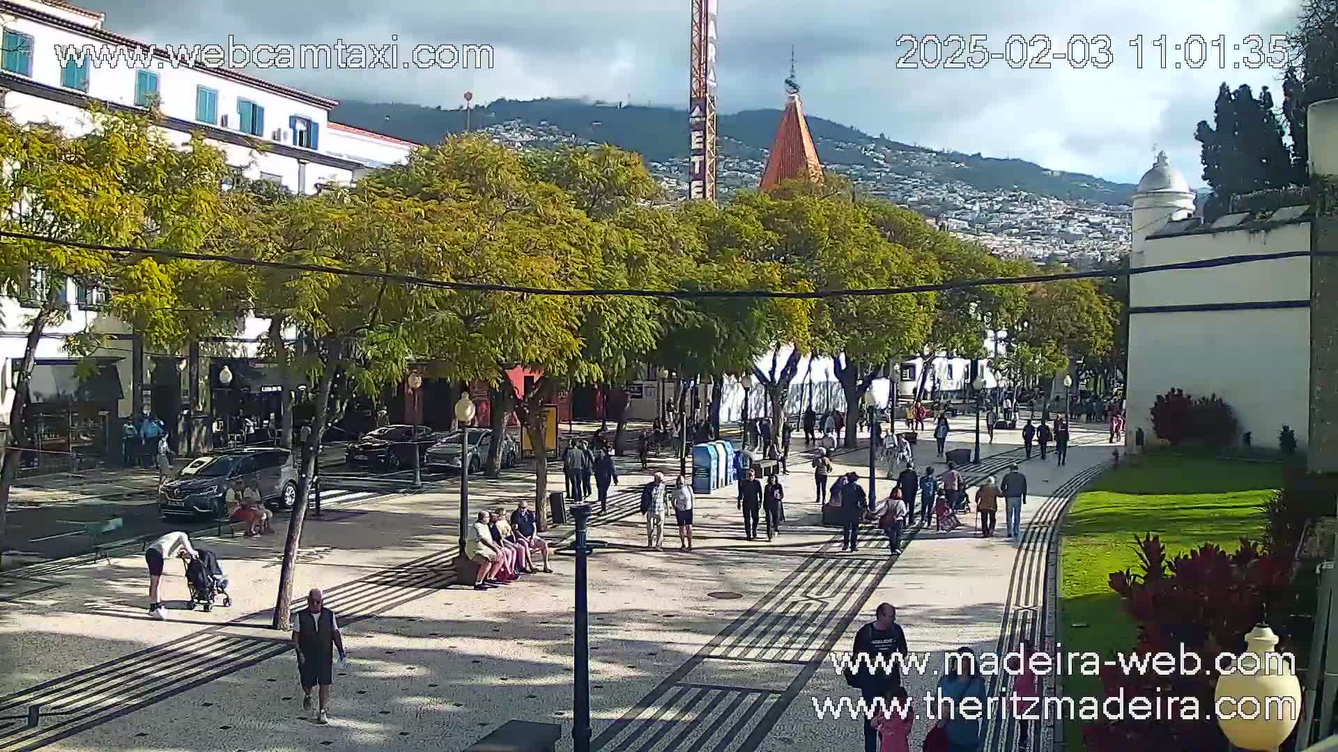 A paved pedestrian area with people walking, sitting, and standing, lined with trees and buildings under a partly cloudy sky.