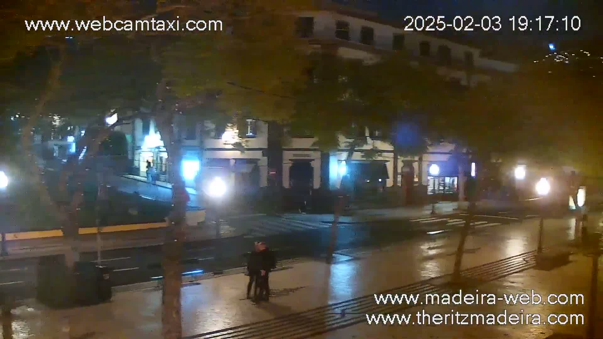 A wet, nighttime street scene shows a multi-story building with streetlights illuminating the sparsely populated street and two people standing near a crosswalk.