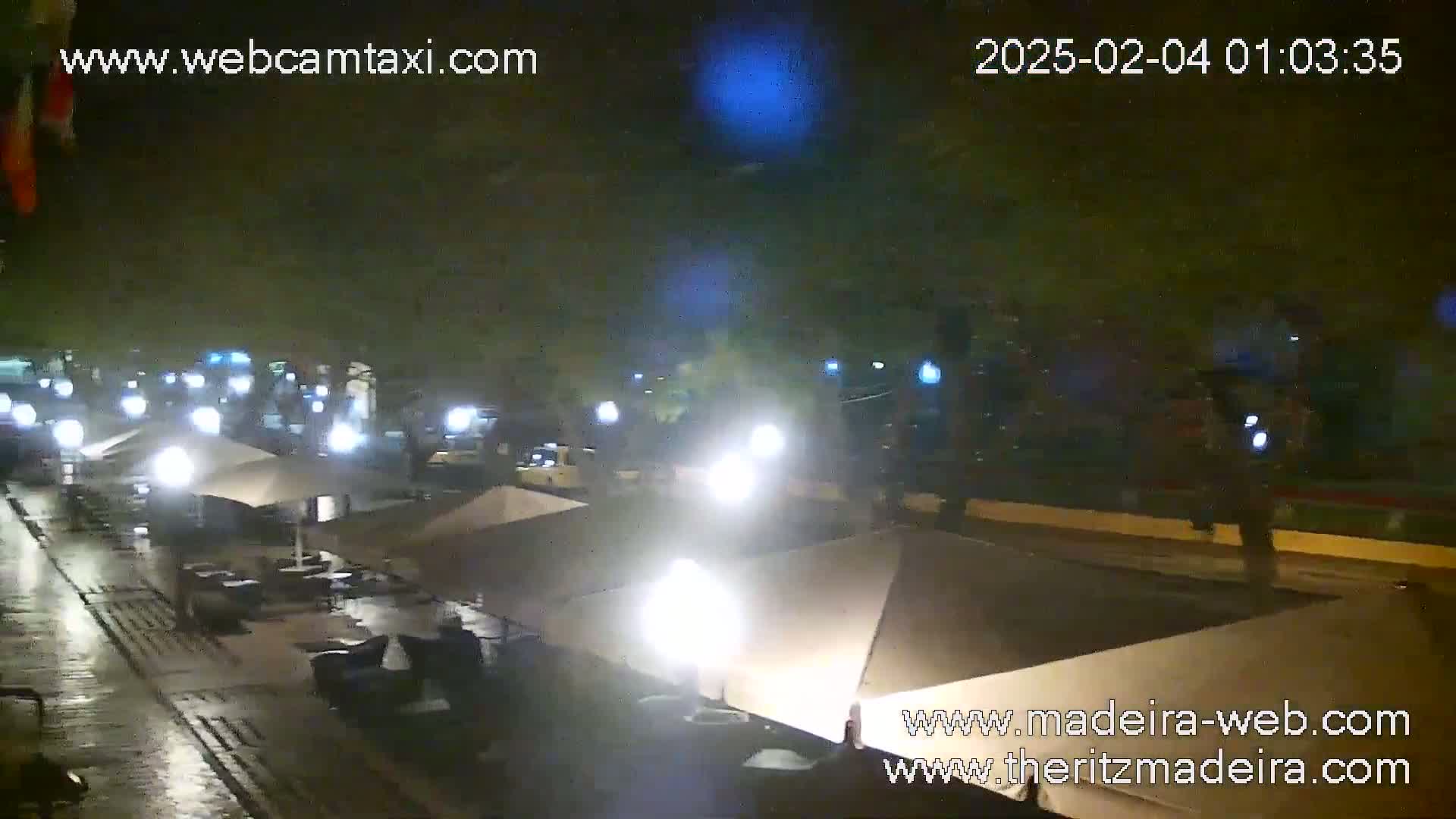 A wet, nighttime street scene shows several closed patio umbrellas and some parked cars under dimly lit streetlights.