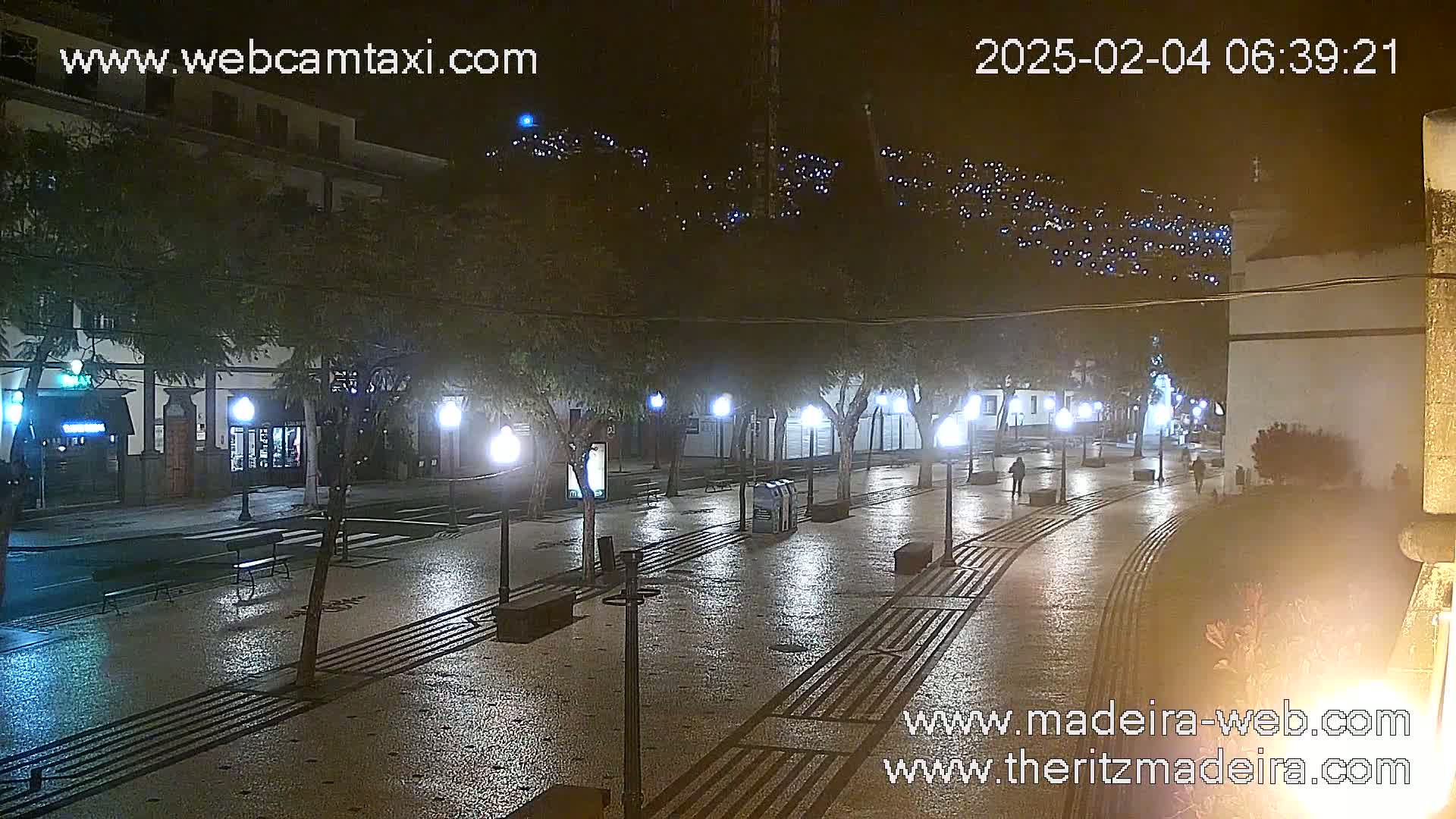 A wet, nighttime street scene shows a paved plaza lined with trees and lampposts, with buildings visible in the background, and a hillside illuminated with lights in the distance.