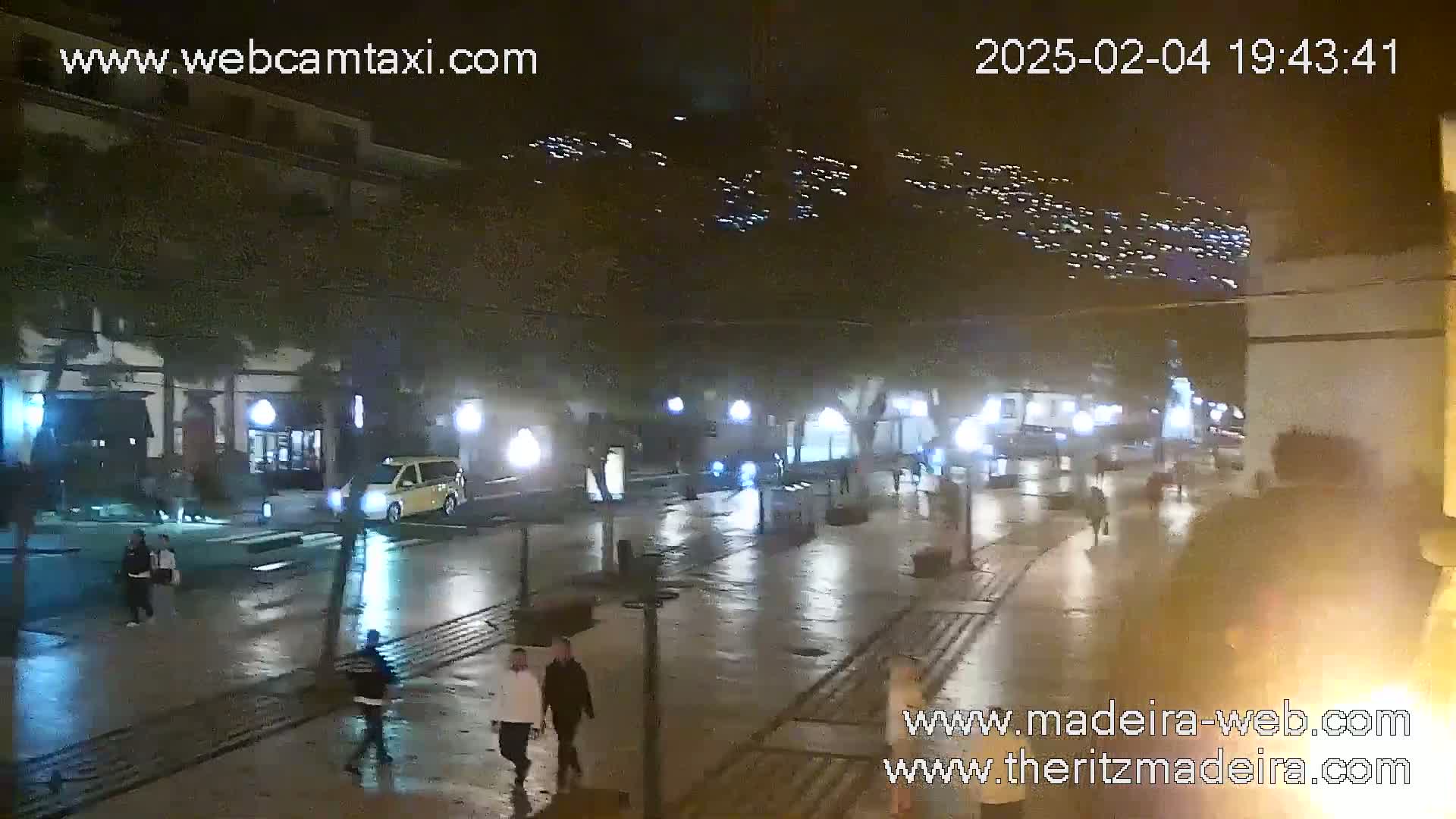 A rain-slicked nighttime street scene shows pedestrians walking past illuminated buildings and shops, with a hillside of lights visible in the distance.