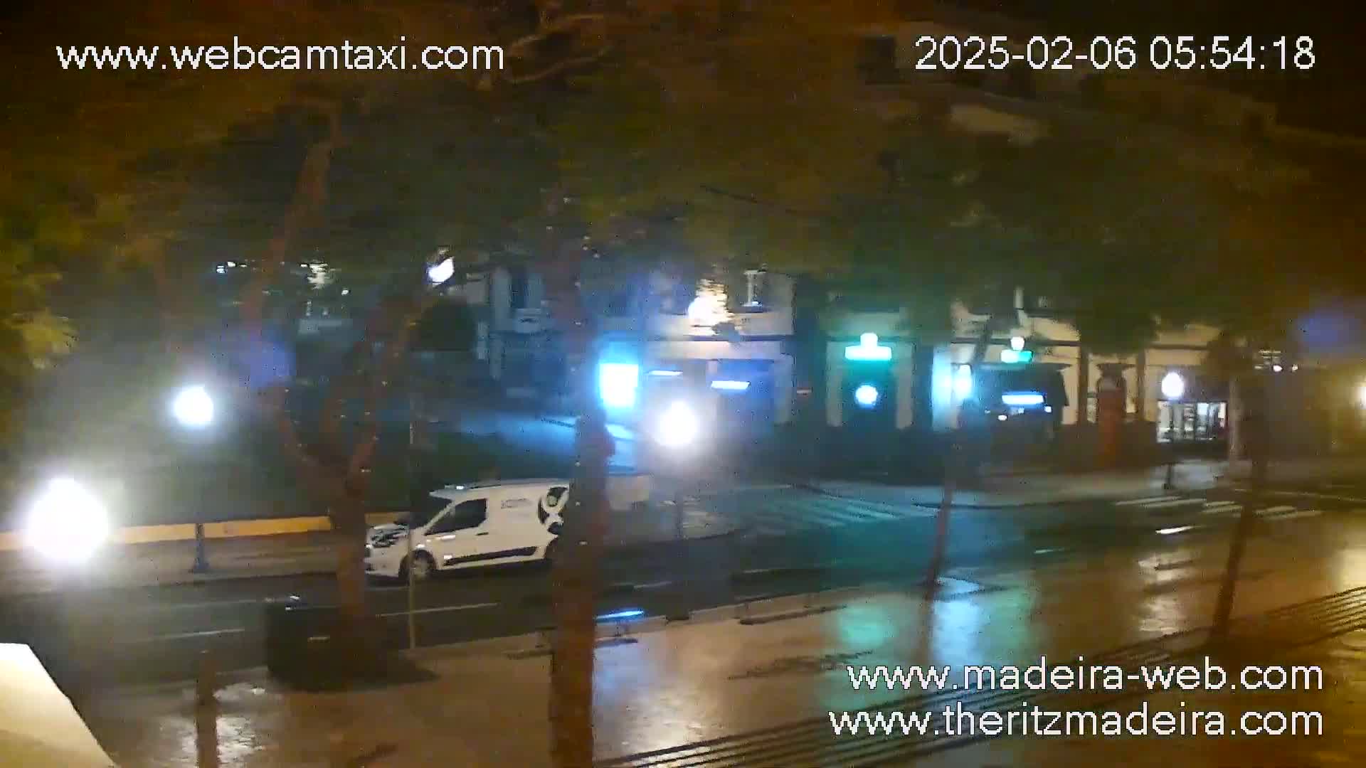 A wet, nighttime street scene shows a white van parked near a building with illuminated signage, under streetlights.