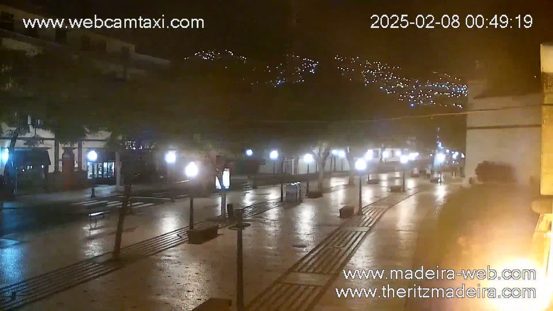 A wet, empty plaza at night is lit by streetlights, with a hillside of lights visible in the distance.