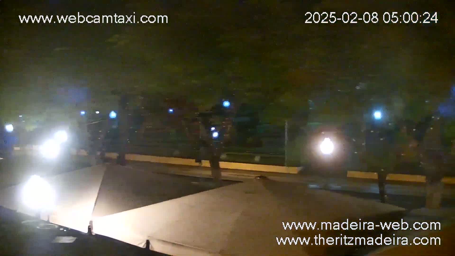 At night, several people walk past closed umbrellas and bright lights on a paved area near some trees.