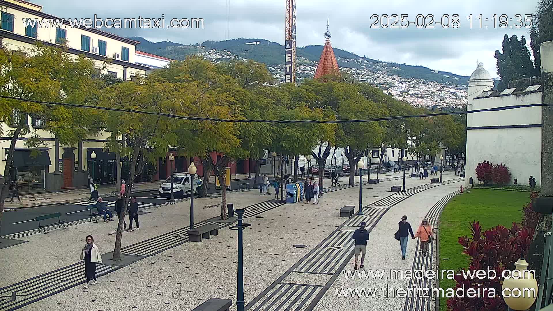 A paved pedestrian walkway lined with trees and buildings, with people walking around under an overcast sky.