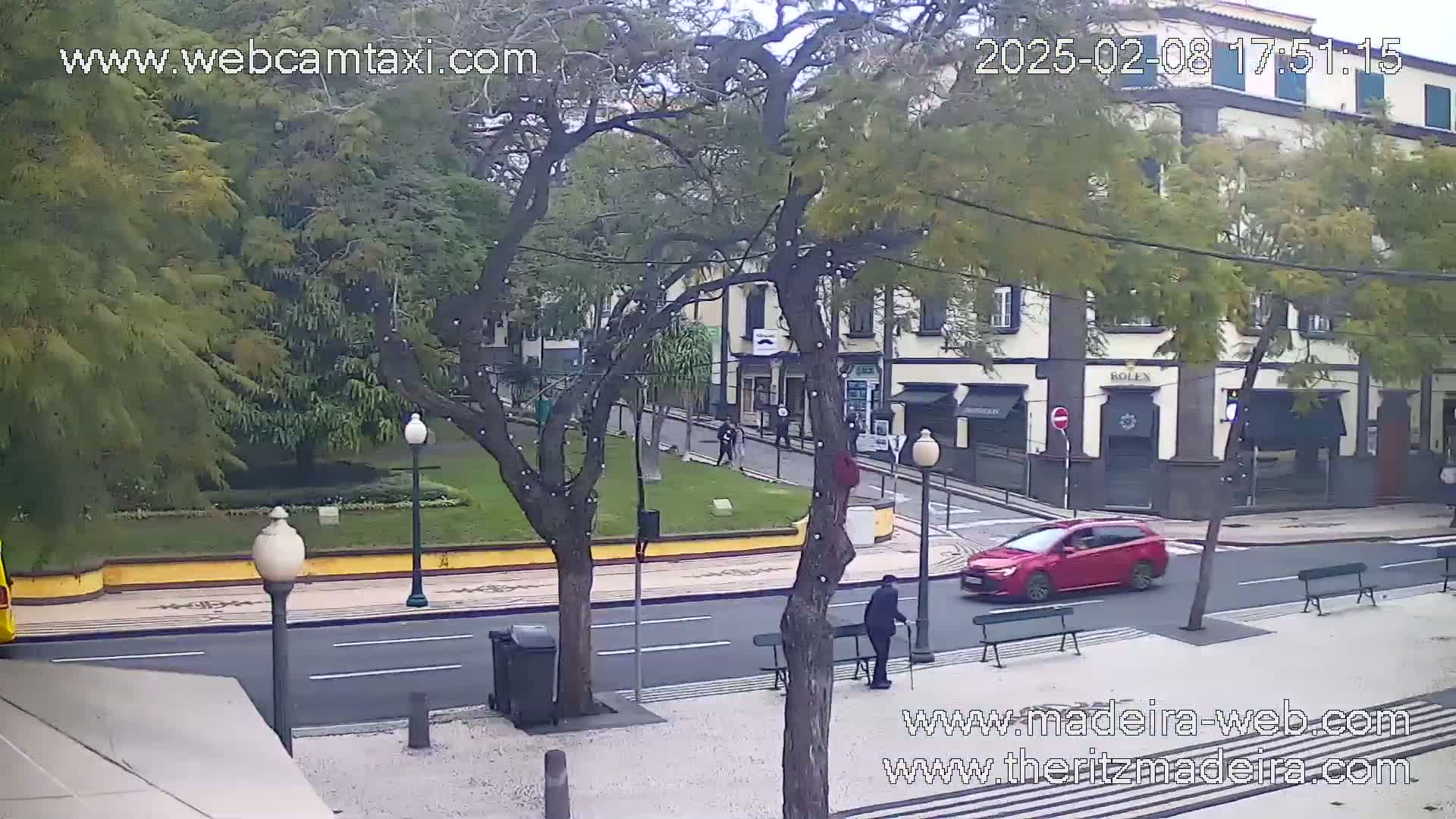 A mostly overcast day shows a street scene with a red car driving past a small park, shops, and pedestrians.
