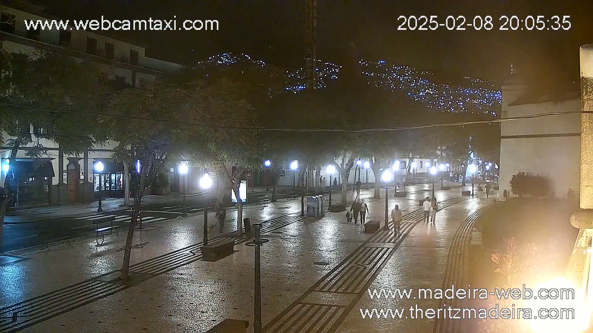A wet, nighttime street scene shows a paved plaza with lampposts, trees, and a few pedestrians walking.