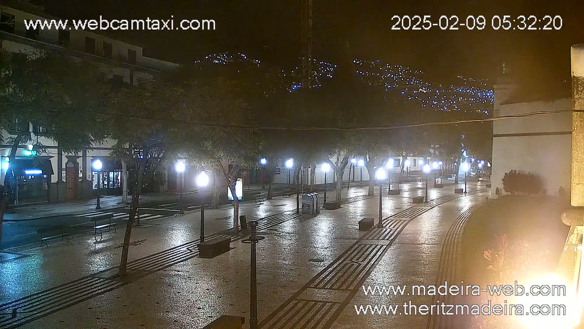 A wet, empty town square at night is illuminated by streetlights, with buildings and hills visible in the background.