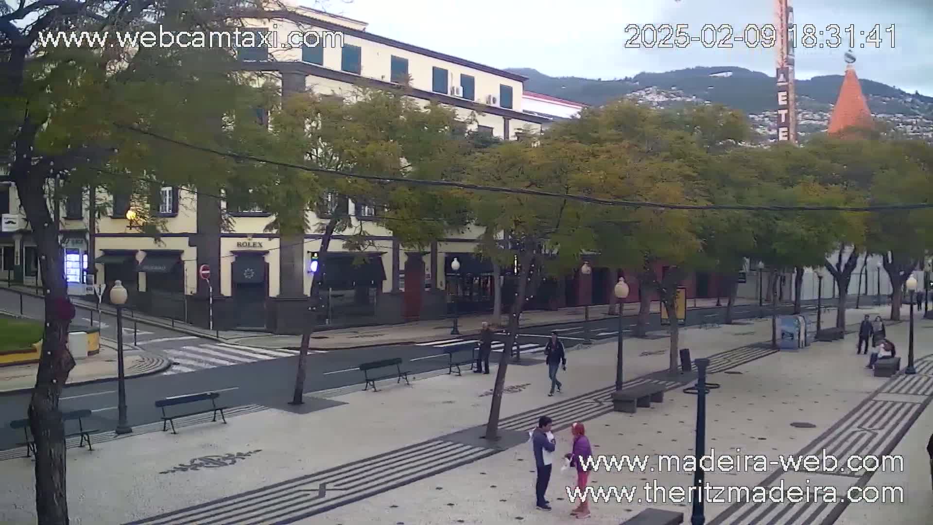 A paved plaza with benches and a few pedestrians, in front of a multi-story building under an overcast sky.