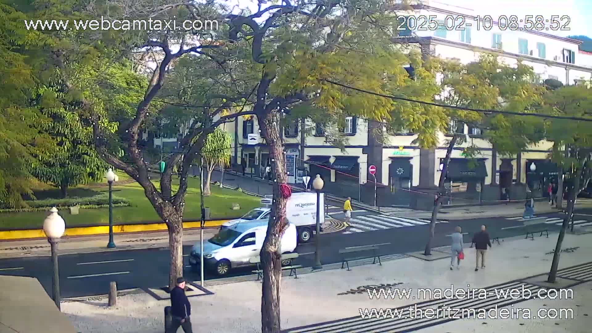 A sunny street scene shows a pedestrian crossing, several people walking, and a few vehicles including a van, parked along the side of the road in a tree-lined area.