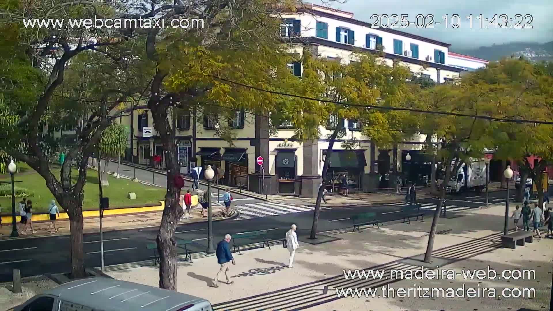 A sunny city street scene shows pedestrians walking past a multi-story building with shops on the ground floor, under the shade of trees.