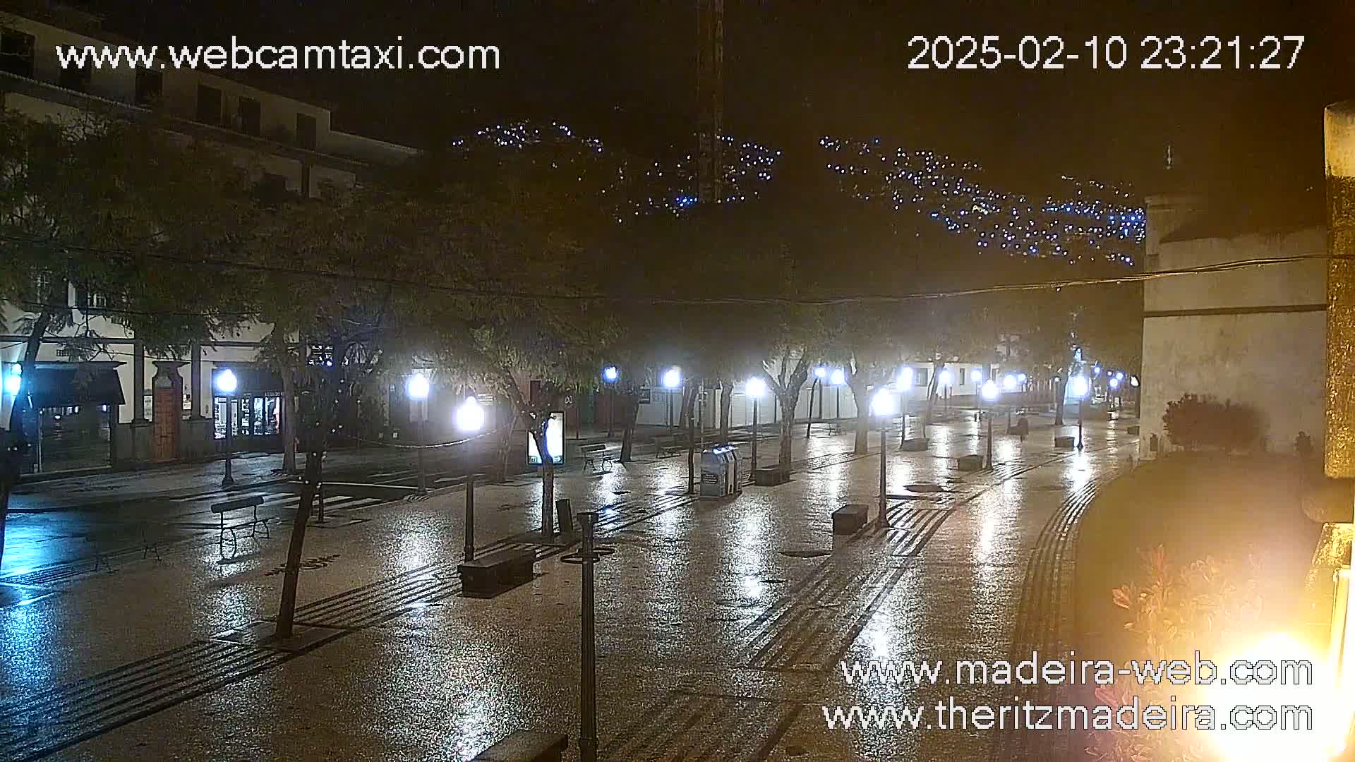 A rain-slicked, nighttime town square is illuminated by streetlights, with buildings lining the sides and hills in the background dotted with lights.