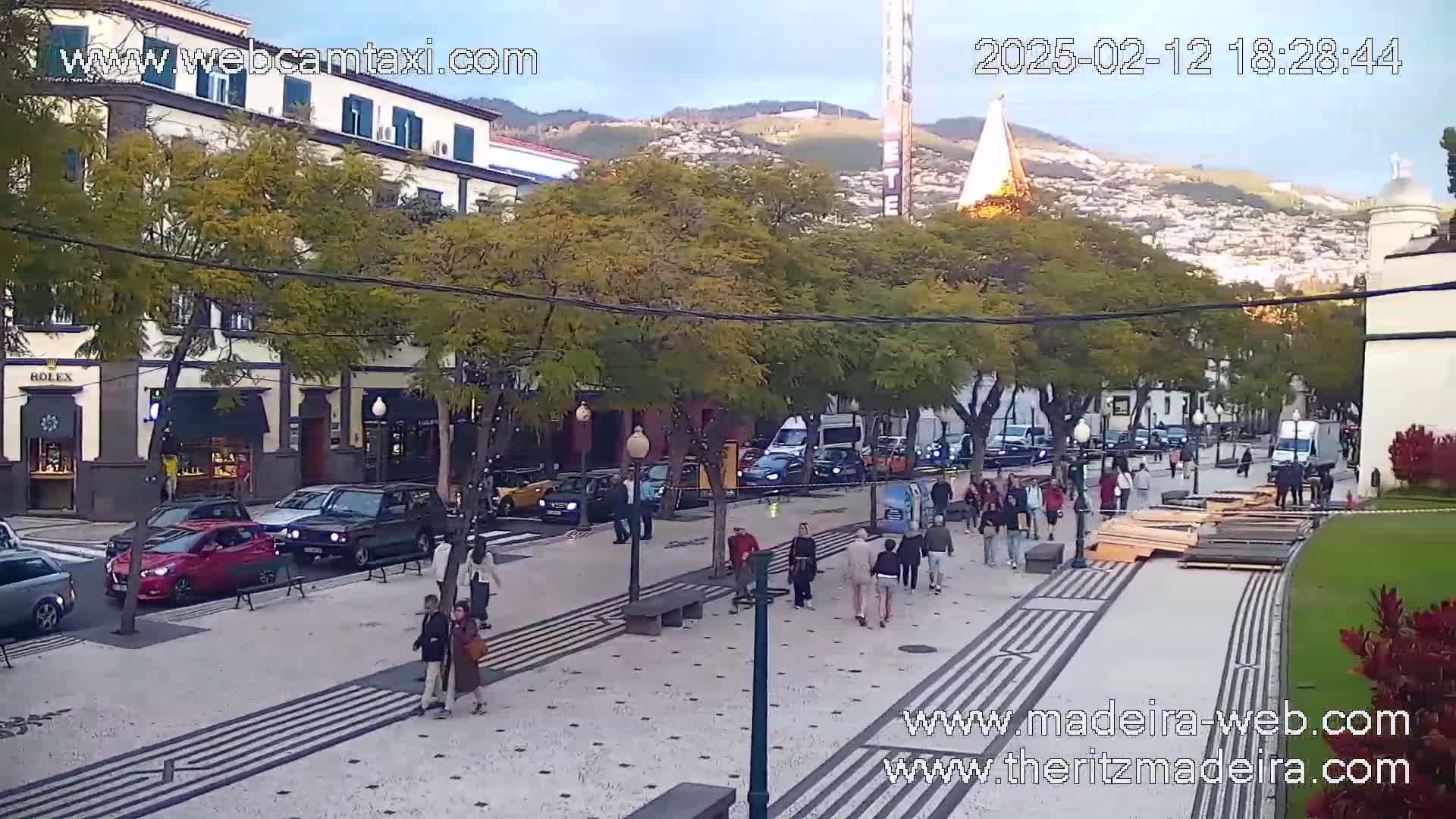 A pedestrianized street scene with cars parked along the side, several people walking, and buildings and trees lining the street under a partly cloudy sky.