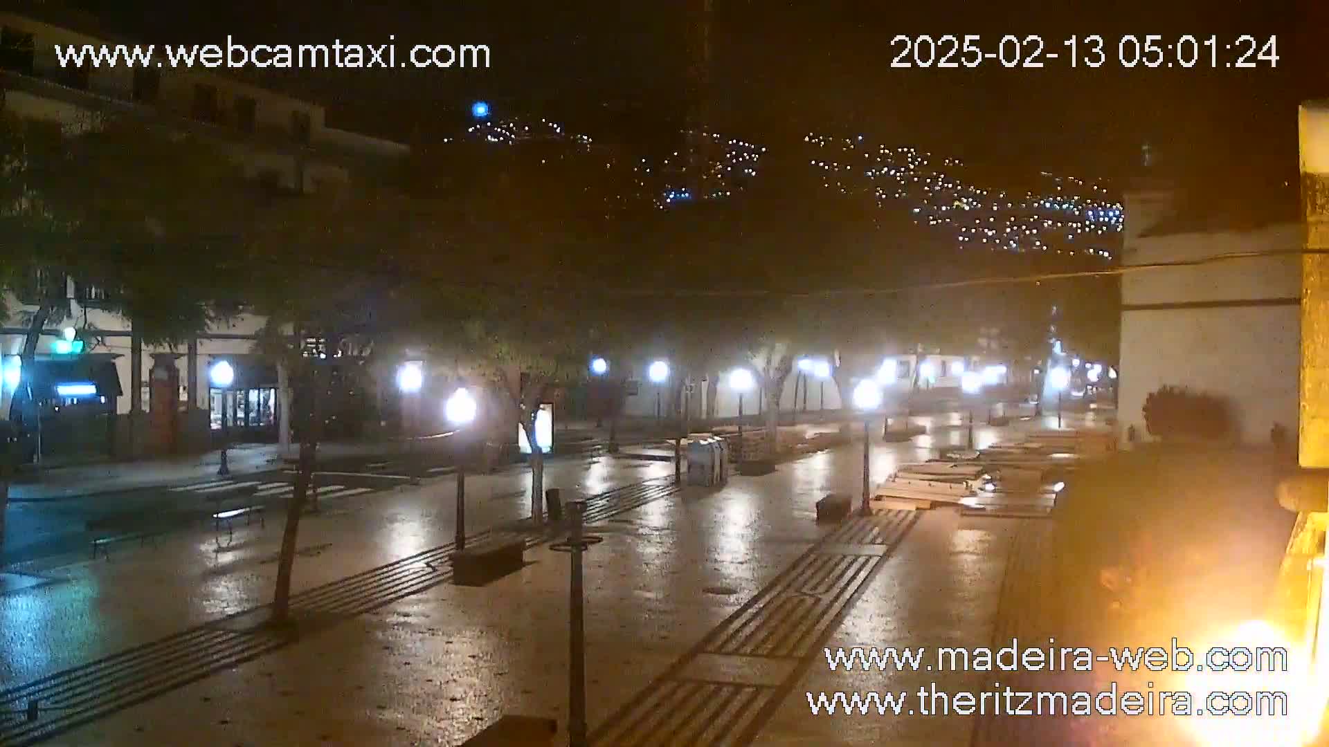 A wet, nighttime street scene shows a town square with streetlights reflecting on a glistening, dark pavement, and distant hillside lights in the background.
