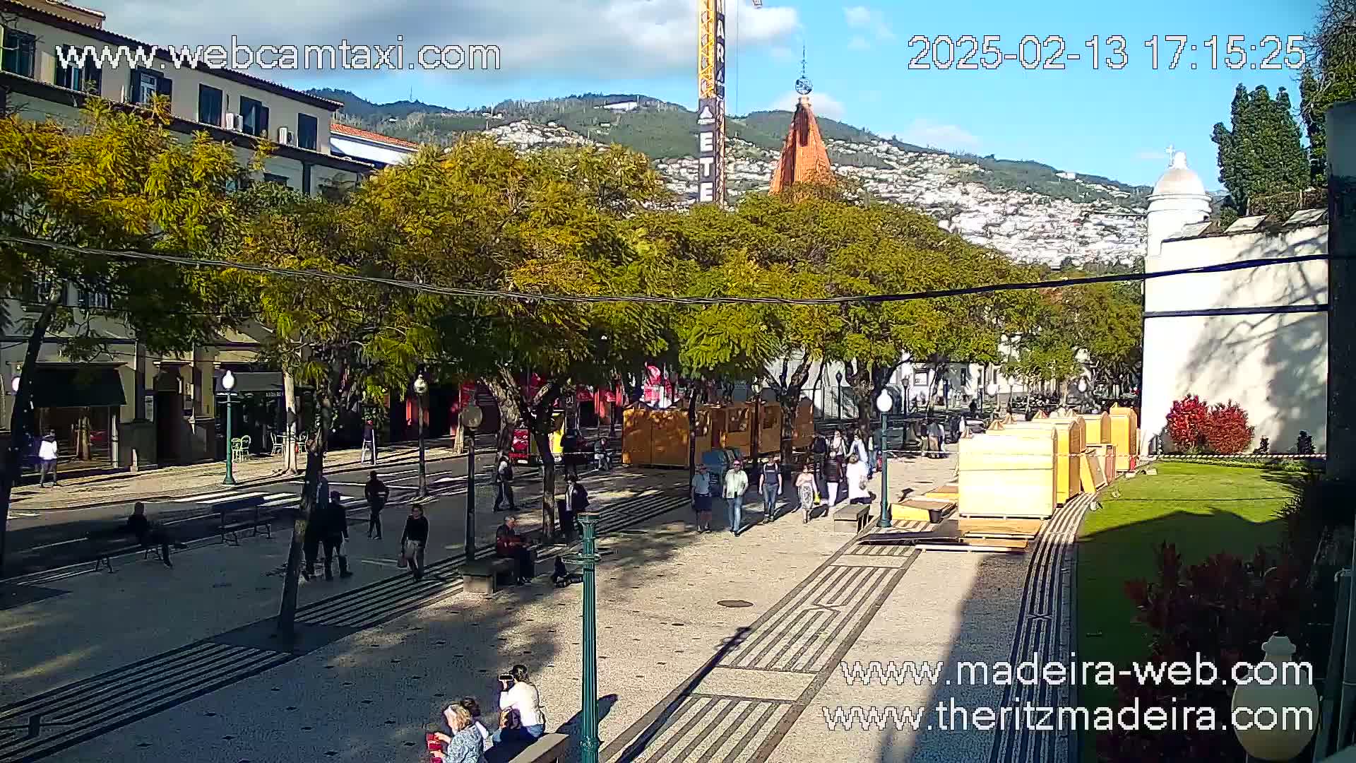 A sunny paved plaza with pedestrians and trees, situated in front of buildings, against a backdrop of hills and a clear sky.