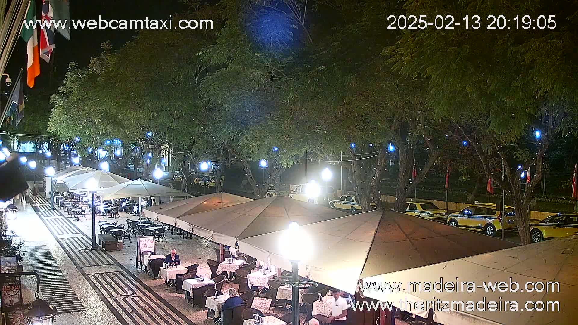 An outdoor nighttime scene shows a paved area with several covered restaurant patios, string lights in trees, and several yellow taxis parked nearby.