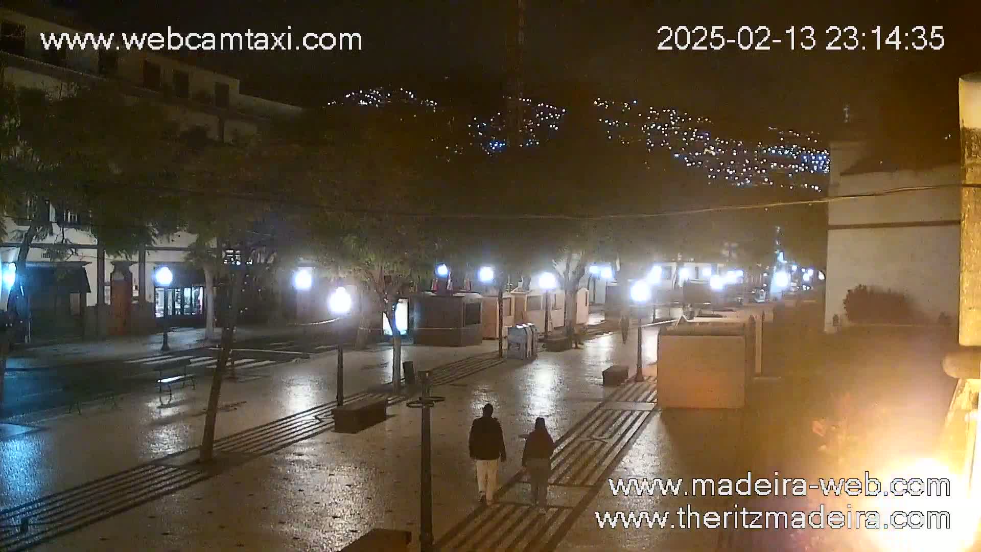 A wet, nighttime street scene shows two pedestrians walking past illuminated buildings and kiosks on a paved plaza, with hillside lights visible in the background.