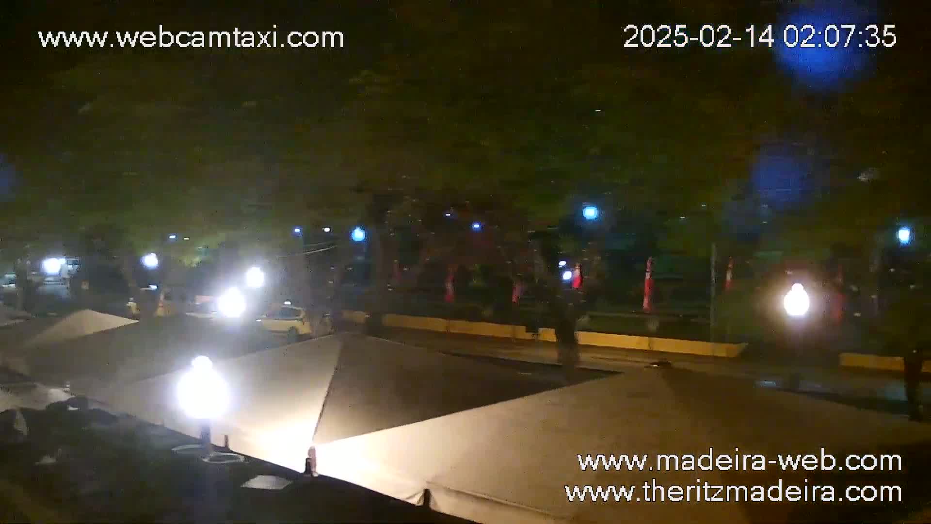A nighttime street scene shows several cars driving past a row of tan canopies under bright lights.