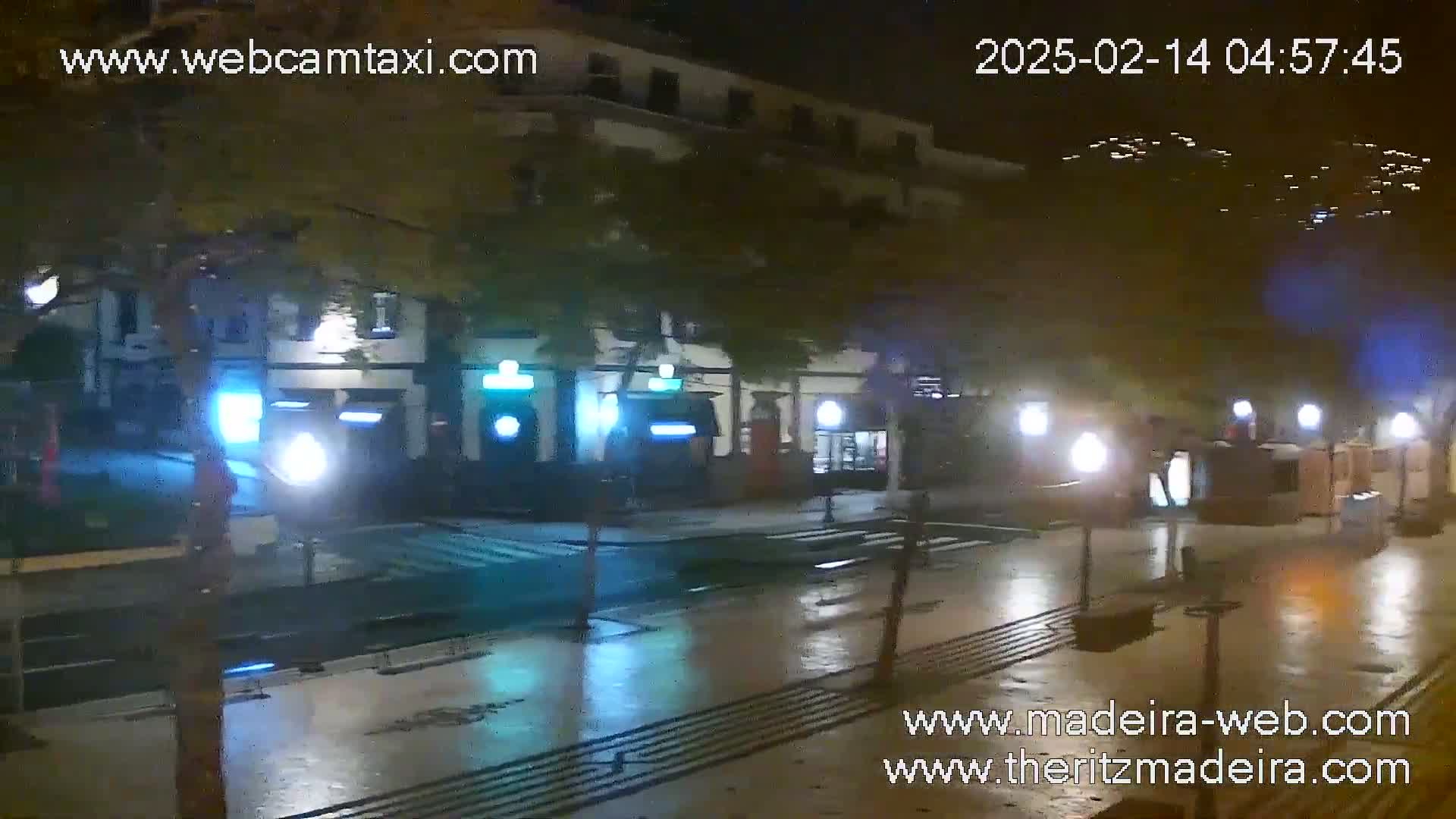 A wet, nighttime street scene shows a row of buildings with illuminated storefronts, streetlights reflecting on the pavement.