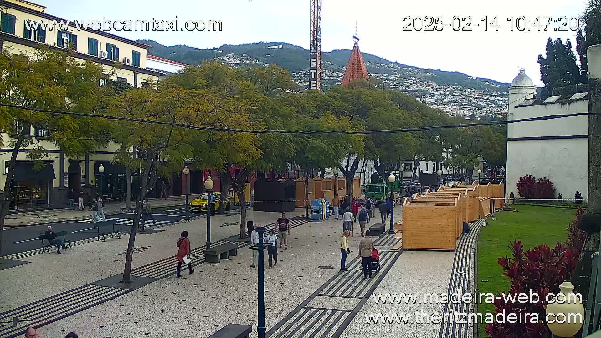 A paved plaza with people walking, some wooden stalls, and several trees under an overcast sky, with buildings and hills in the background.