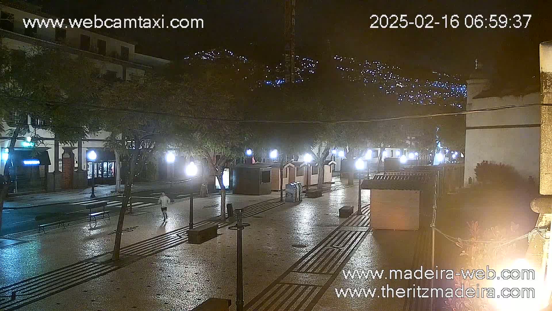 A nighttime view of a wet, paved plaza with small kiosks, streetlights, and a few people, overlooked by buildings on a hillside twinkling with lights.