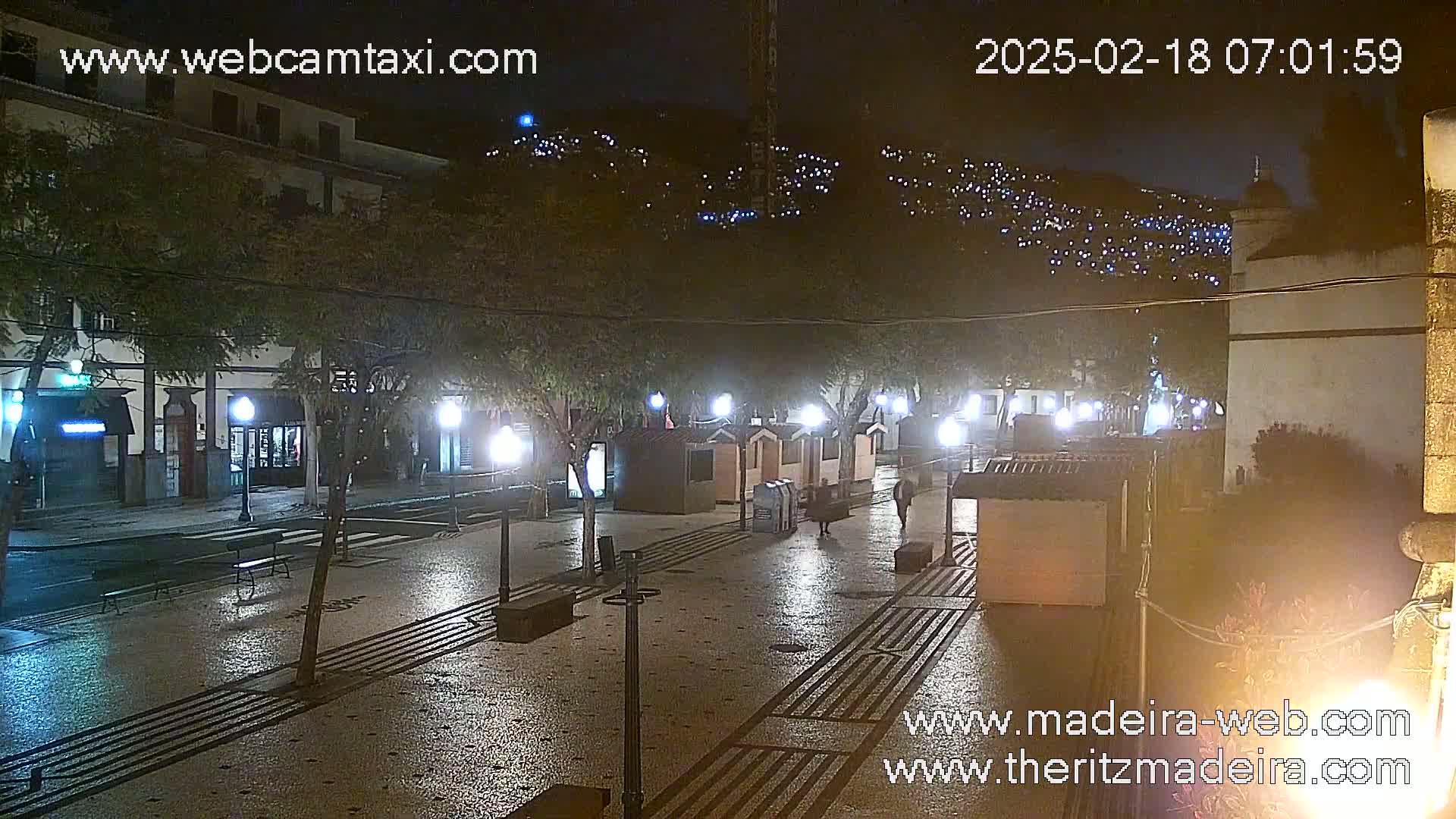 A wet, nighttime street scene shows a plaza with small buildings, trees, and lights, all under a dark sky illuminated by distant lights on a hill.