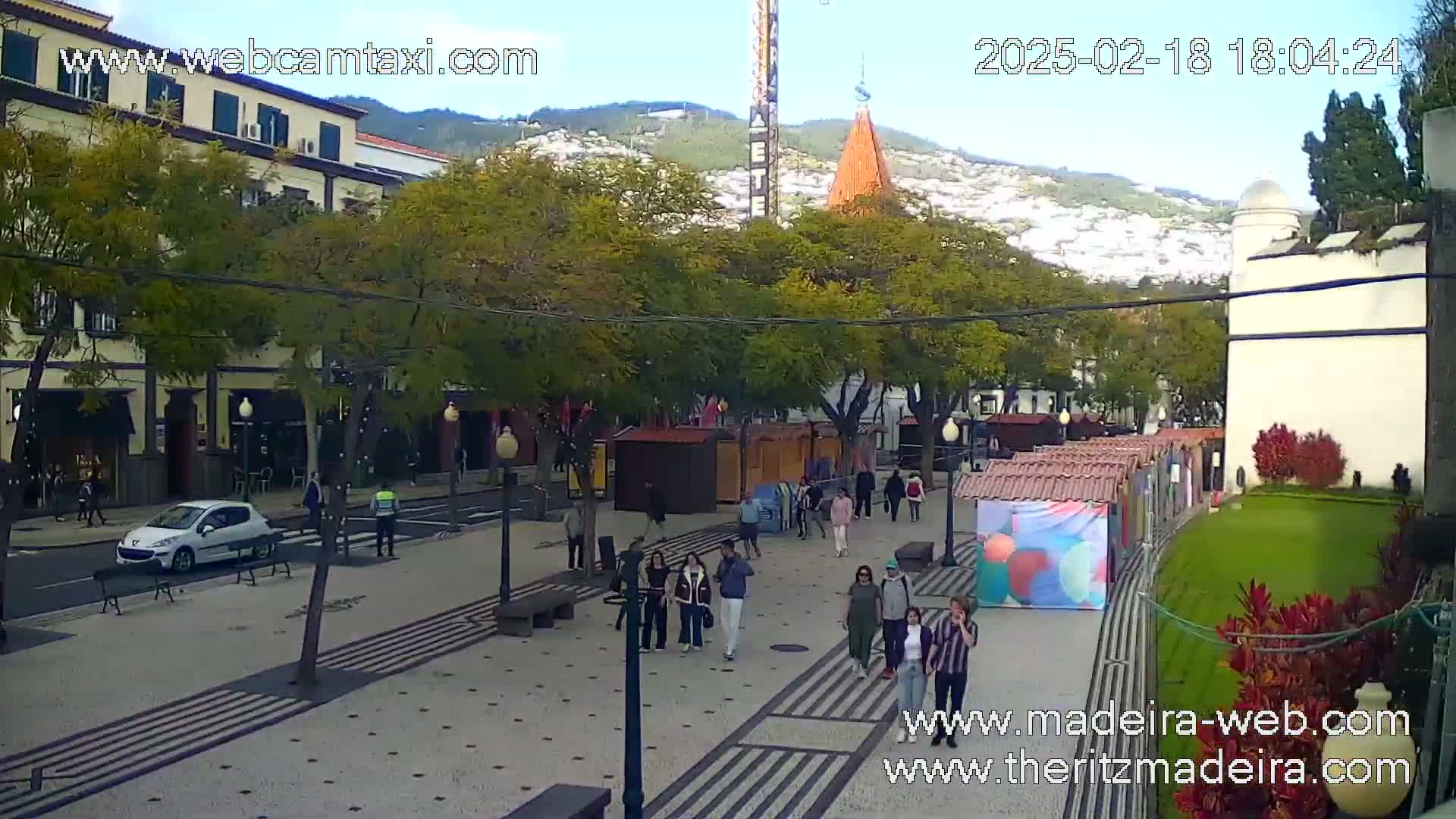 A paved plaza with people walking, small market stalls, and buildings, under a mostly clear sky.