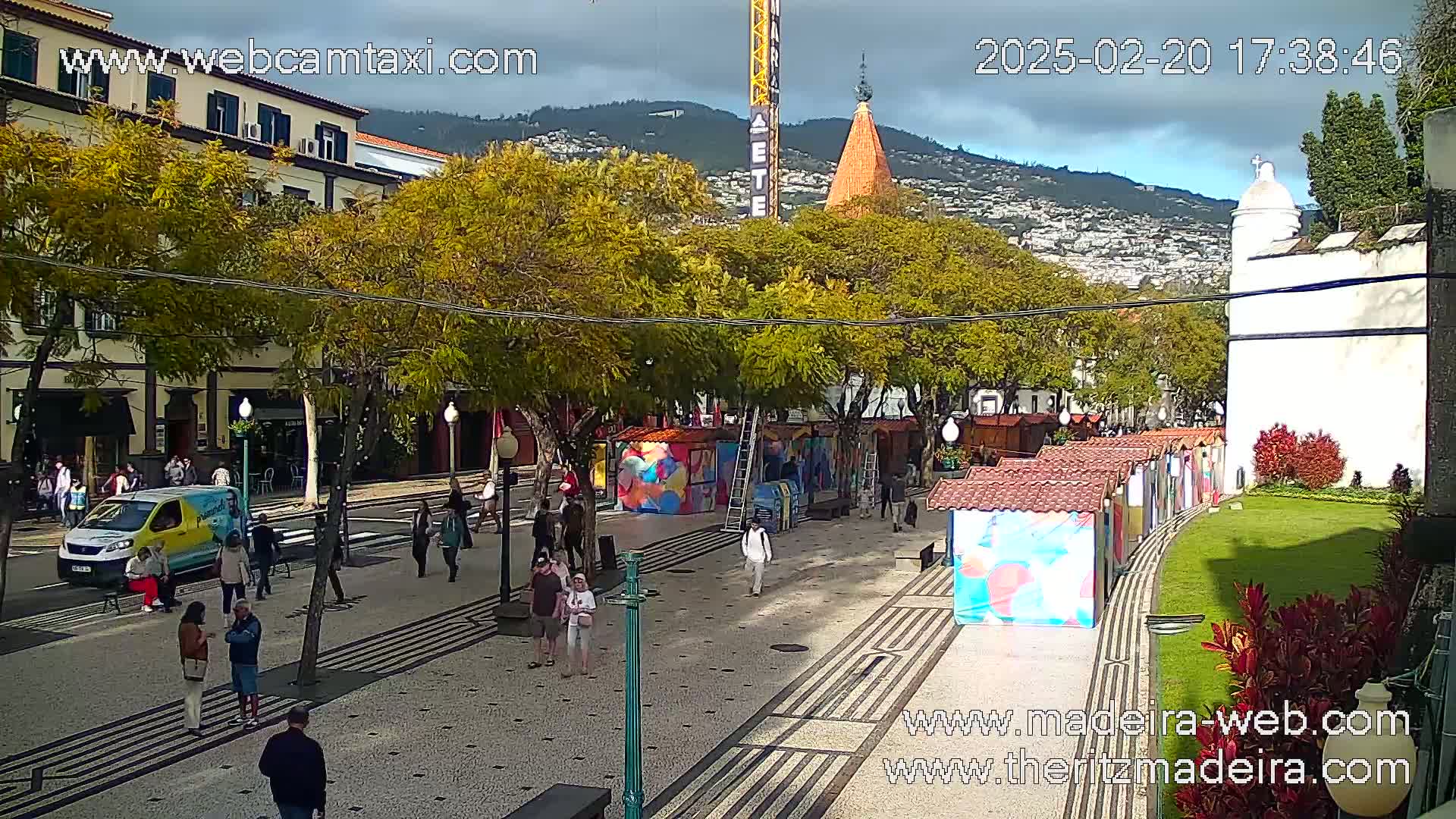 A paved pedestrian area with colorful stalls and trees, under a cloudy sky, features people walking and a van parked nearby, with a mountain backdrop.