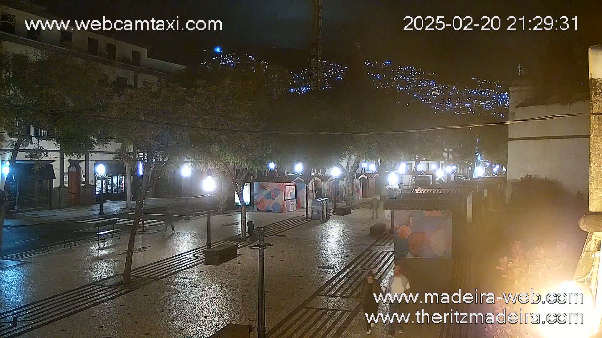A nighttime view of a town square with illuminated lampposts and small kiosks, overlooking a hillside dotted with lights under a clear, dark sky.