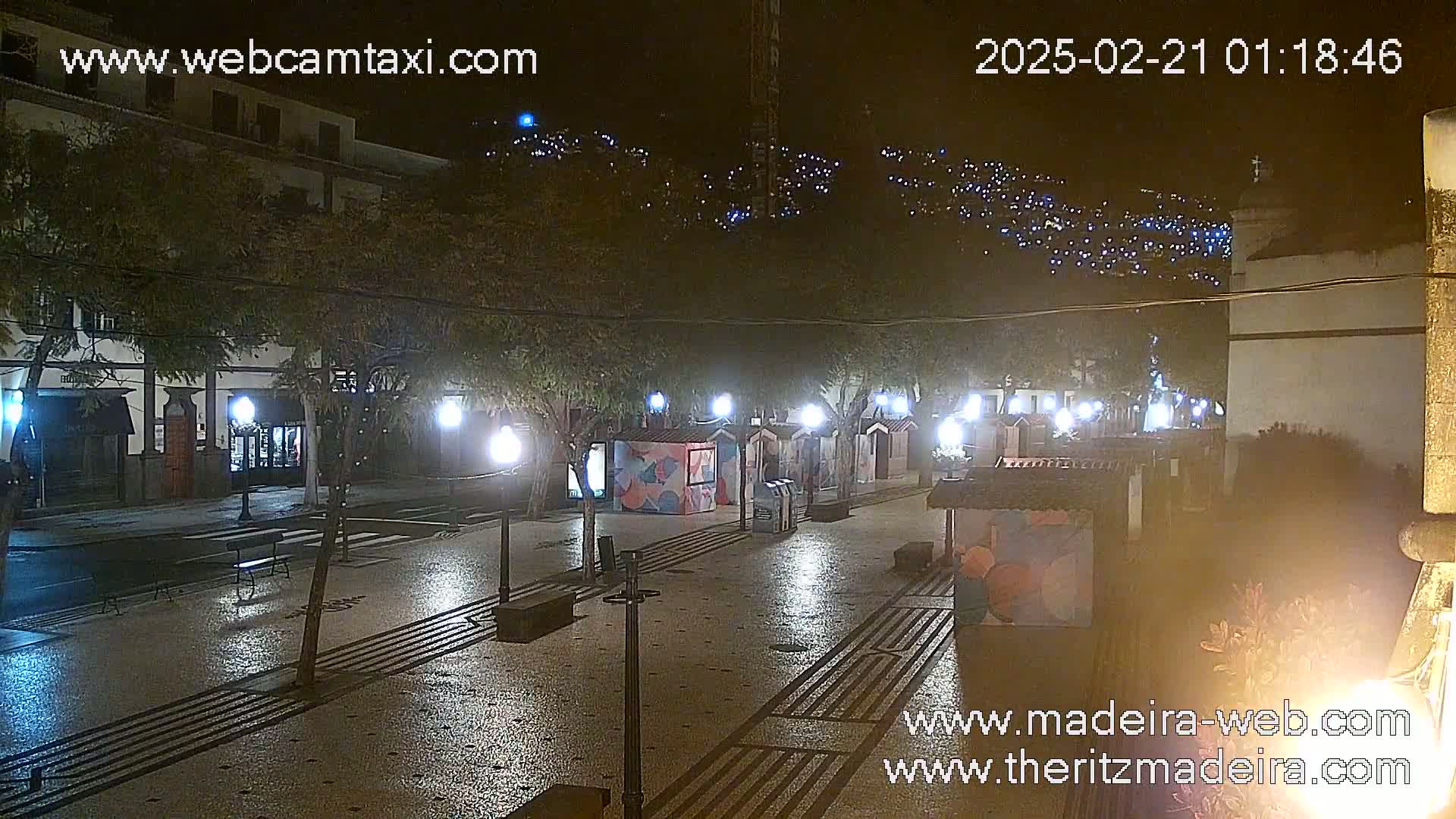 A nighttime view of a town square with several small, brightly lit stalls and streetlights, surrounded by trees and buildings.