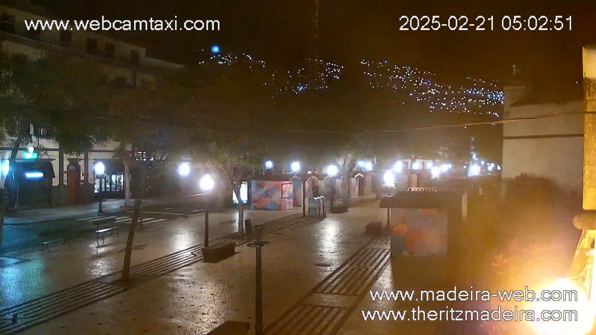 A wet, nighttime town square is illuminated by streetlights and the lights of small buildings and stalls, with a hillside dotted with lights visible in the background.