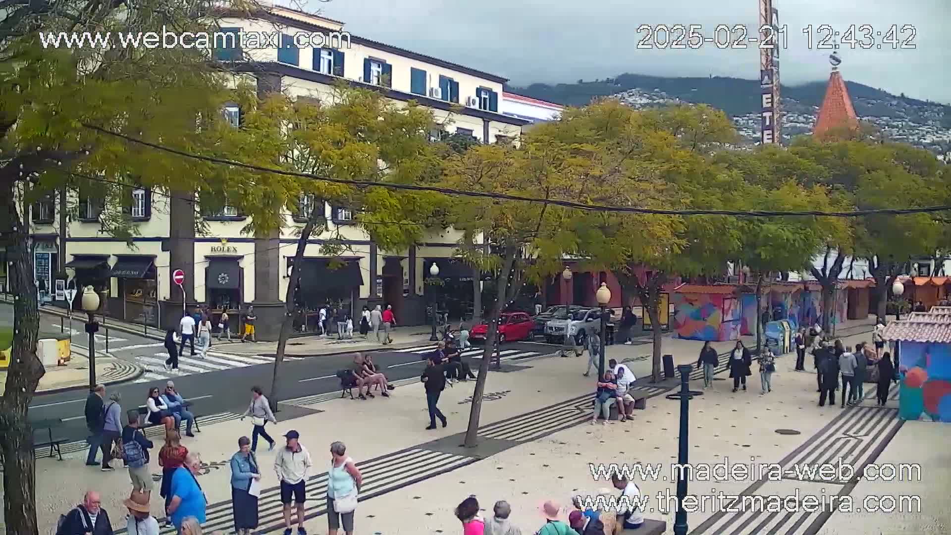 A city street scene on a partly cloudy day shows people walking and sitting on benches in front of buildings and shops, with cars parked along the street and a mountain visible in the background.