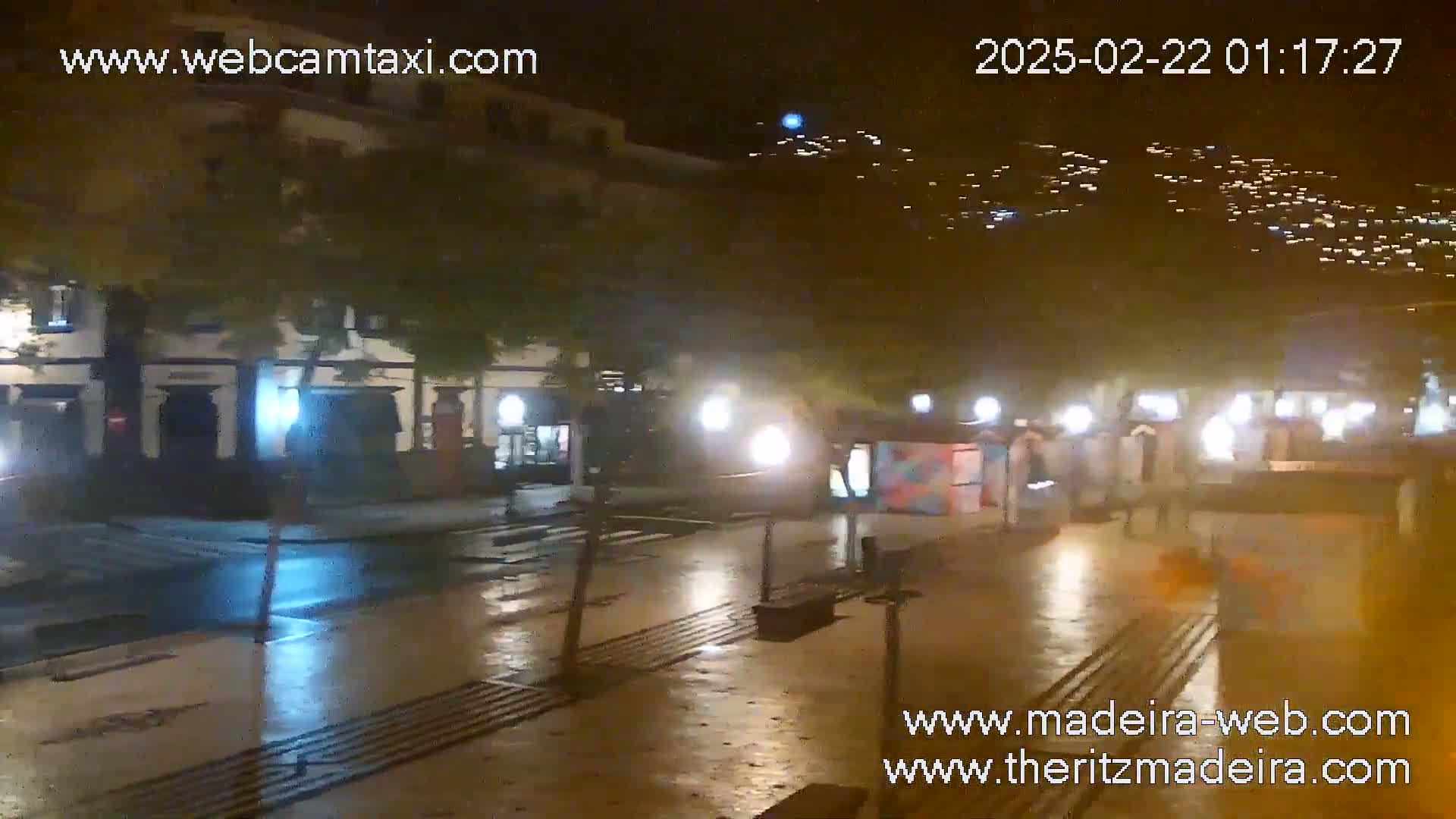A wet, nighttime street scene shows a town square with buildings, streetlights, and a few pedestrians.
