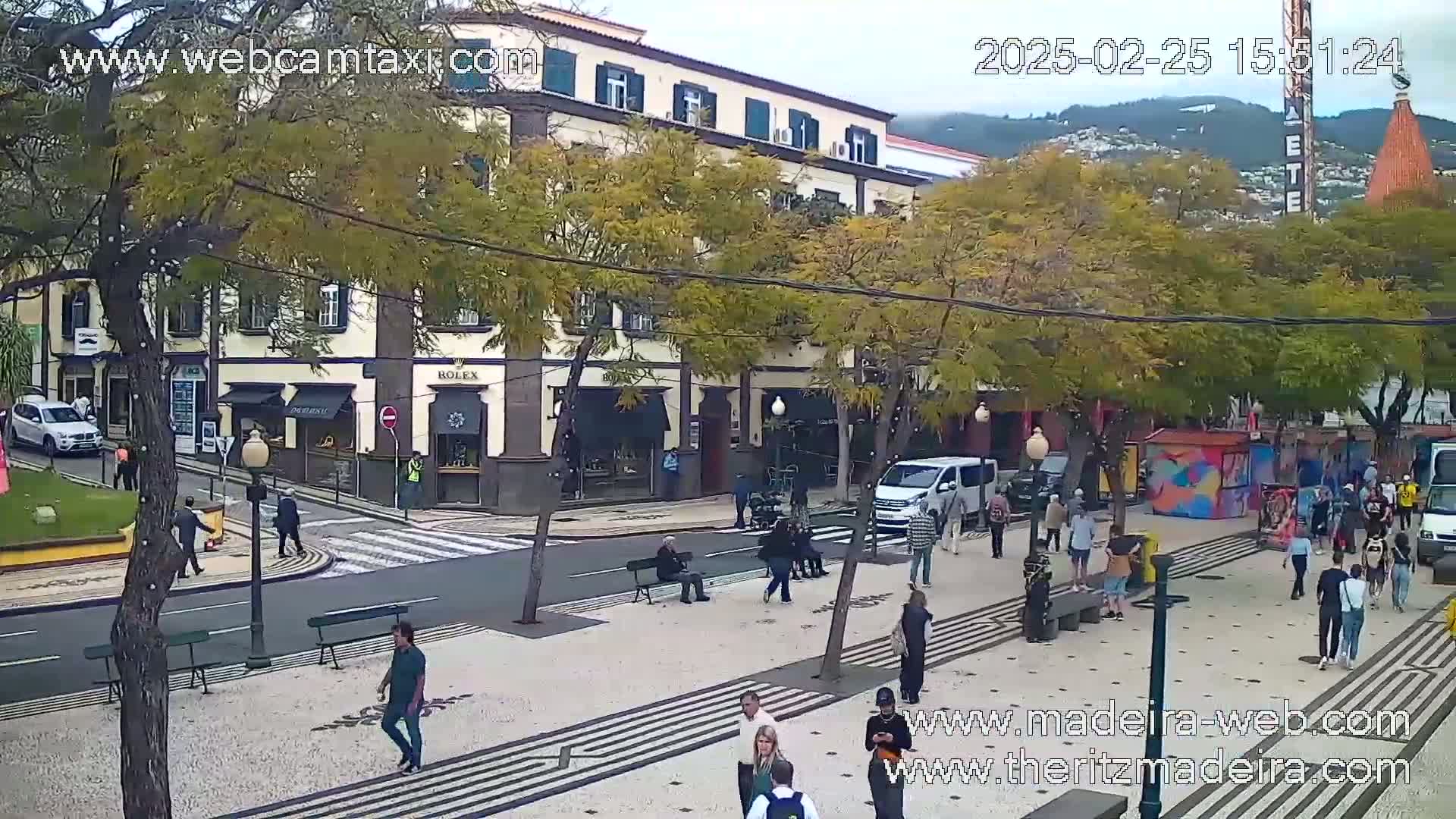 A paved plaza with pedestrians and parked cars is situated in front of a multi-story building under an overcast sky.