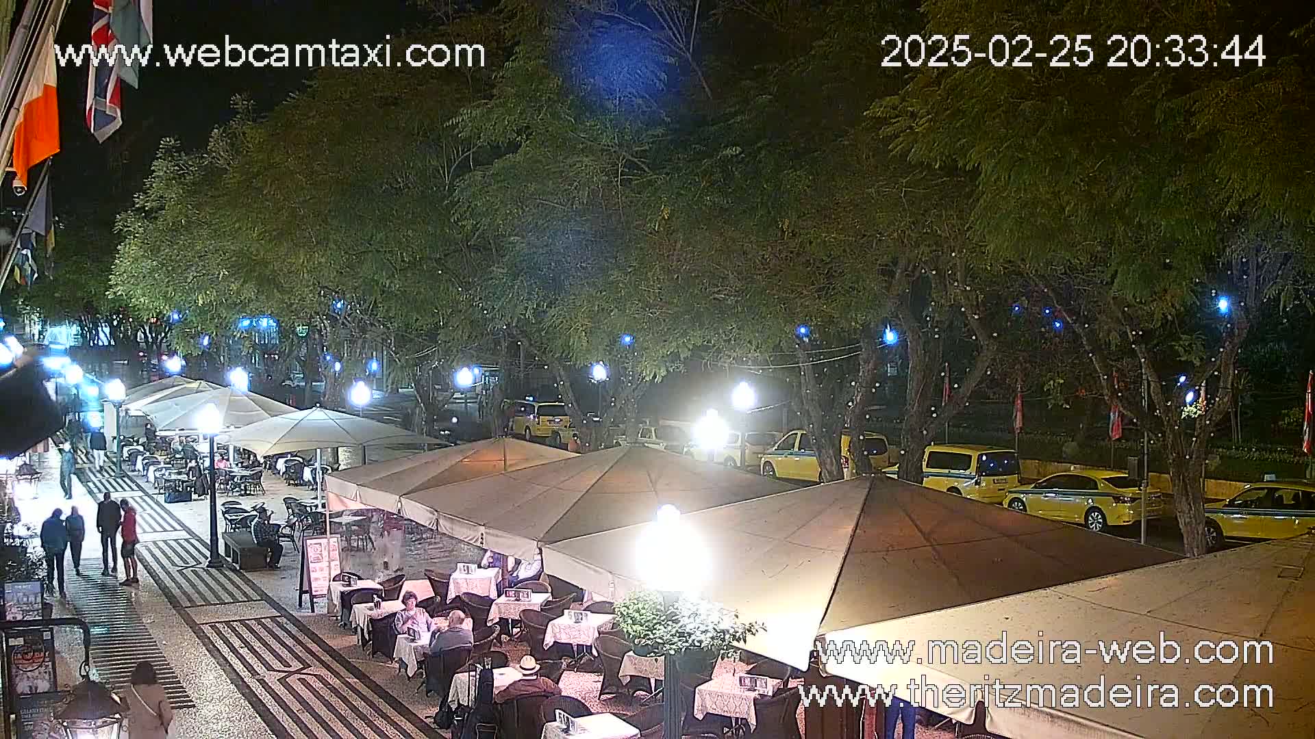 A nighttime outdoor scene shows a paved area with several restaurant tables under beige umbrellas, lined with yellow taxis parked along the side, under leafy trees with blue string lights.
