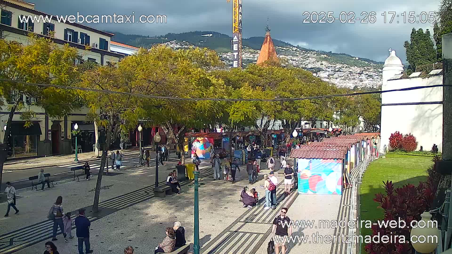 A partly cloudy day reveals a pedestrian street lined with trees, shops, and market stalls, with people walking along a patterned walkway, and hills in the background.