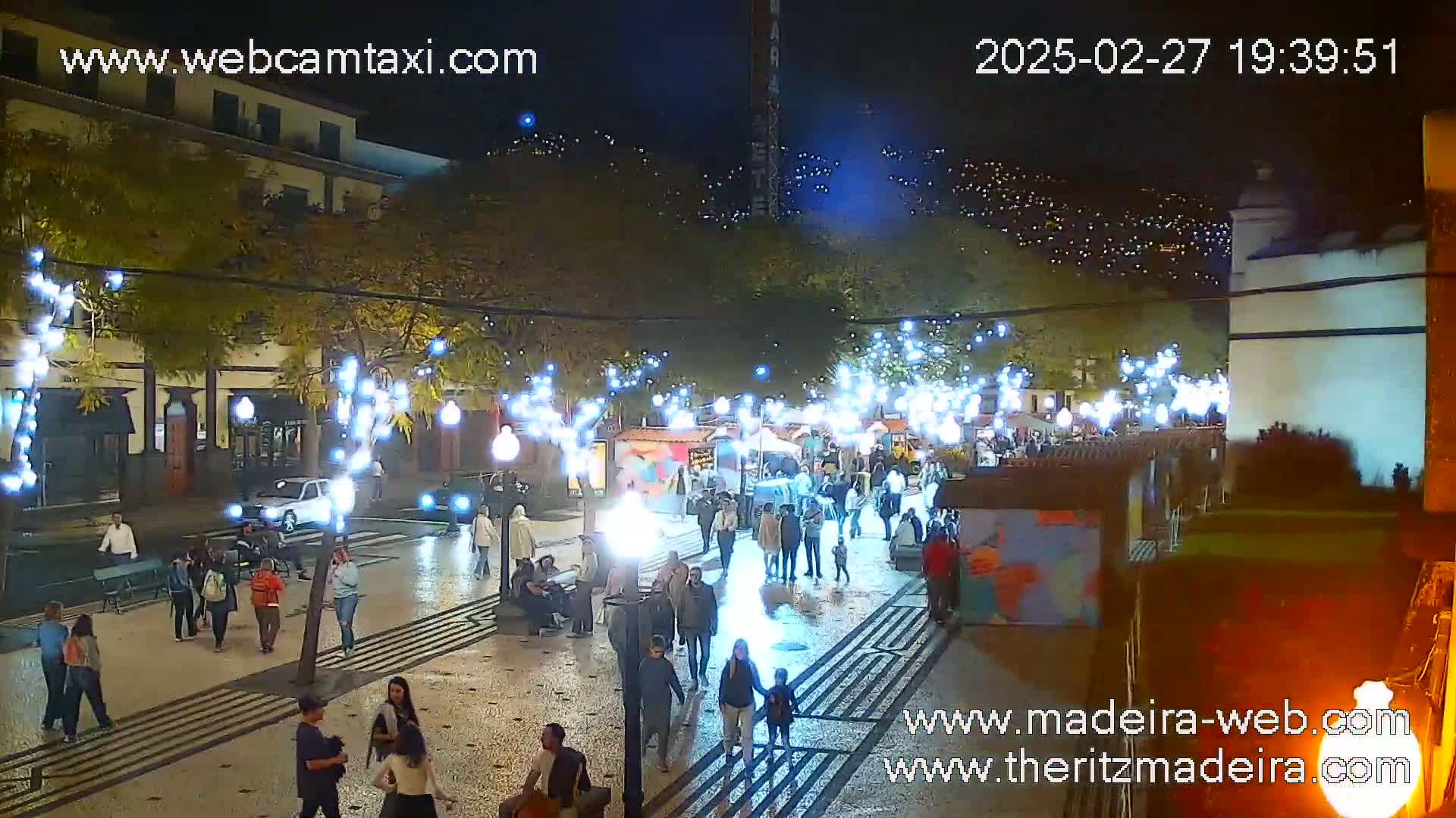 A nighttime scene of a pedestrian plaza with string lights, numerous people walking, and several small market stalls.