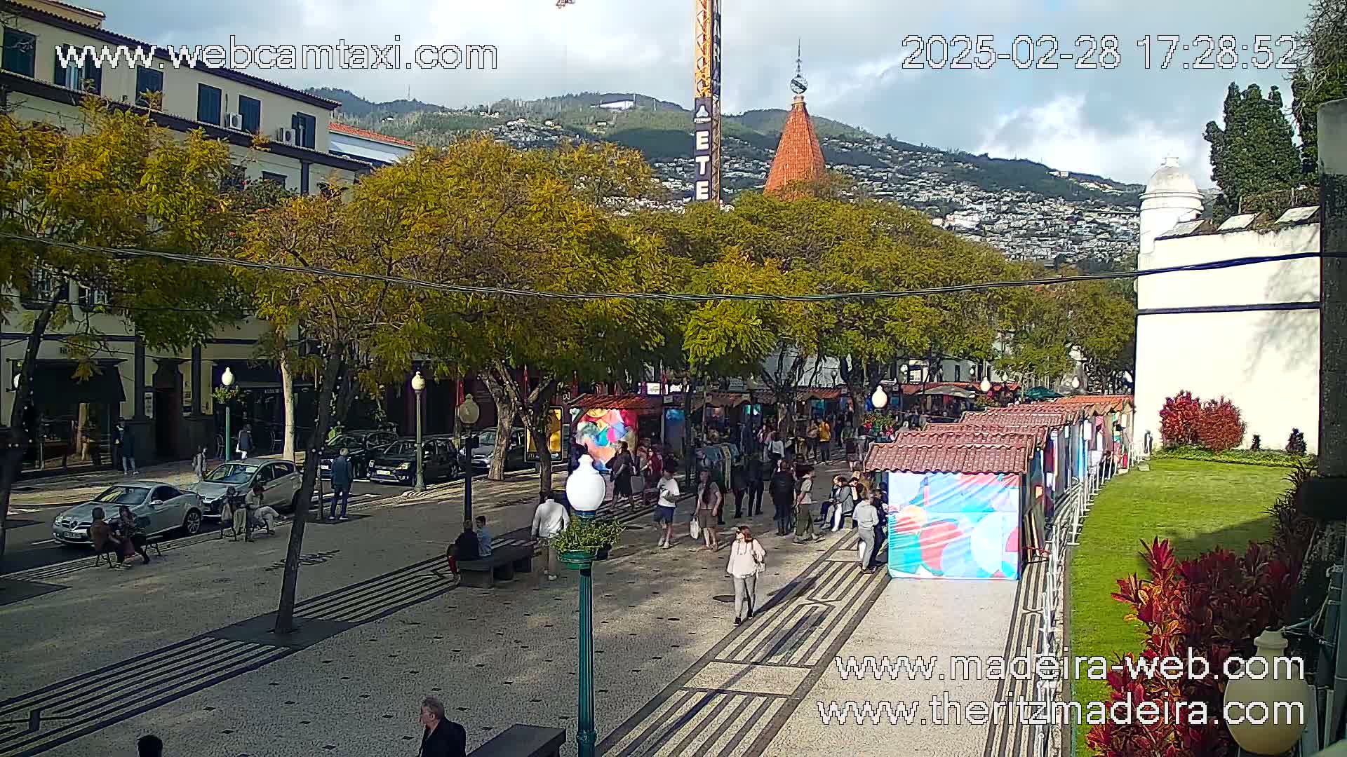 A partly cloudy day reveals a pedestrianized street lined with trees, buildings, parked cars, and market stalls, with people strolling and sitting.