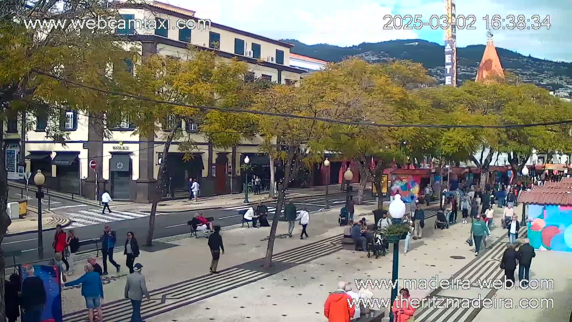 A sunny day on a pedestrianized street lined with trees and buildings, where many people are walking and sitting.