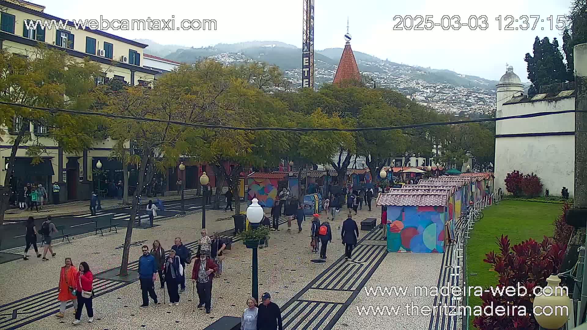 A paved pedestrian area with people walking, lined with trees and small colorful market stalls, under an overcast sky, with hills and buildings visible in the background.