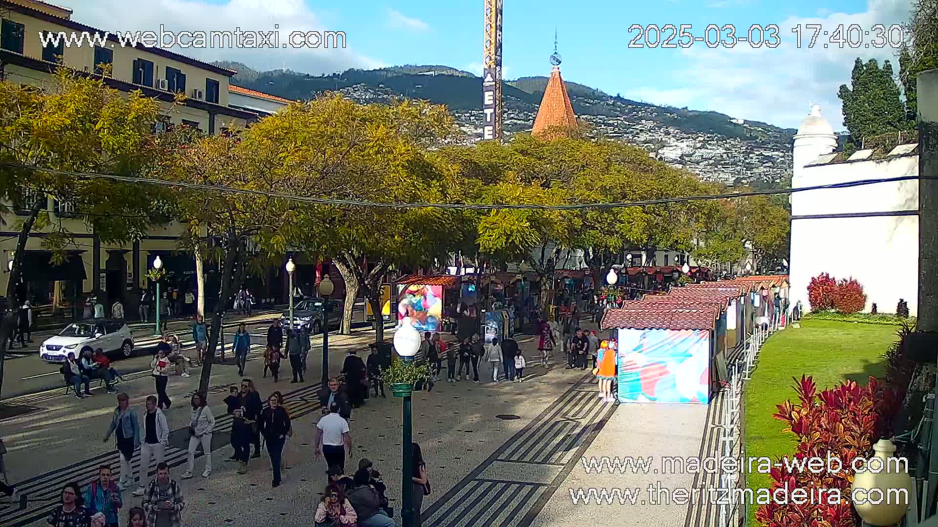 A sunny day reveals a pedestrian street lined with trees, shops, and market stalls, with people walking and cars parked along the side, against a backdrop of mountains.